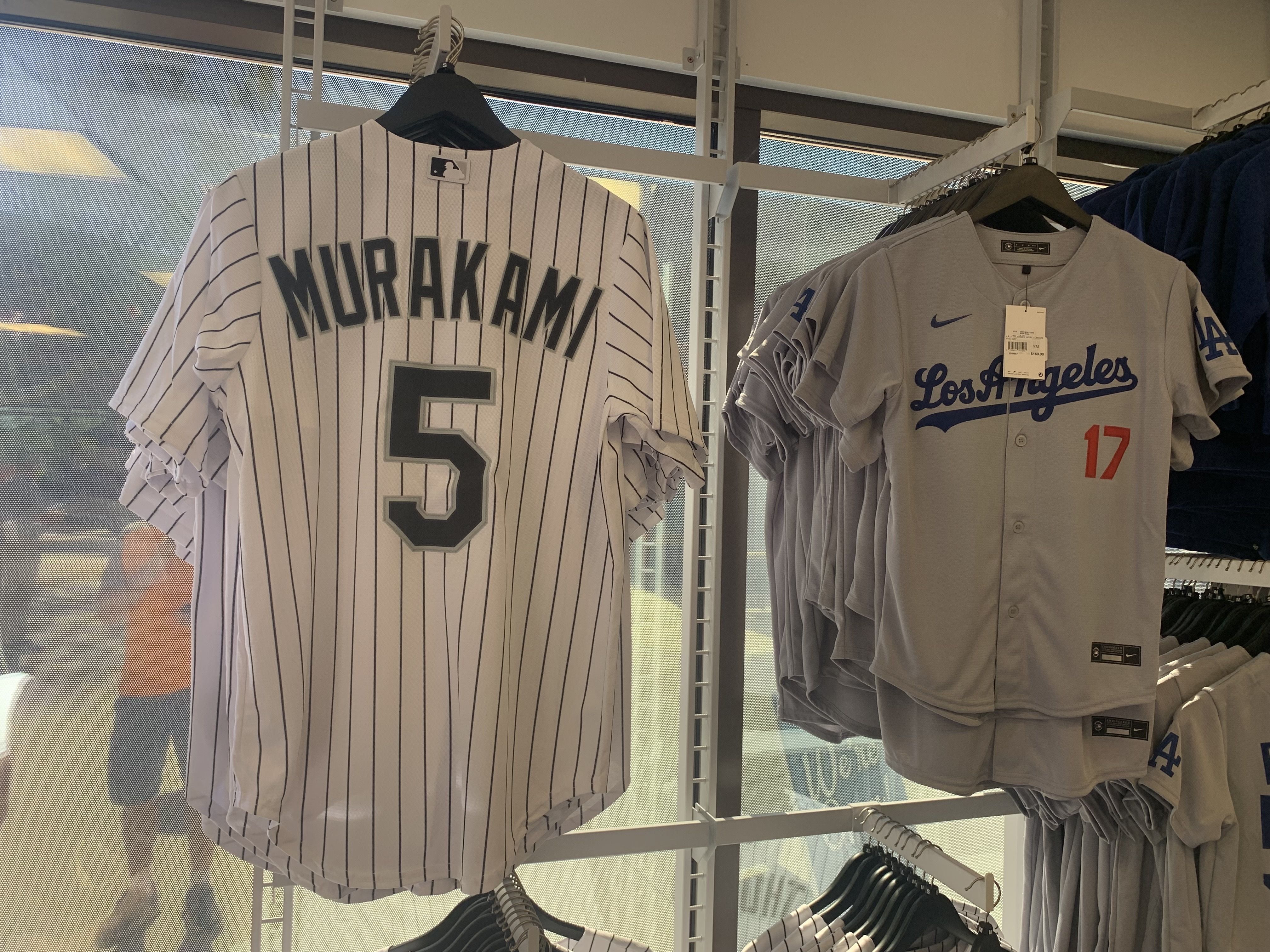 White baseball jersey with black pinstripes and "MURAKAMI 5" on back hanging next to gray Los Angeles Dodgers jersey with blue text and number 17 in red for sale in a store.