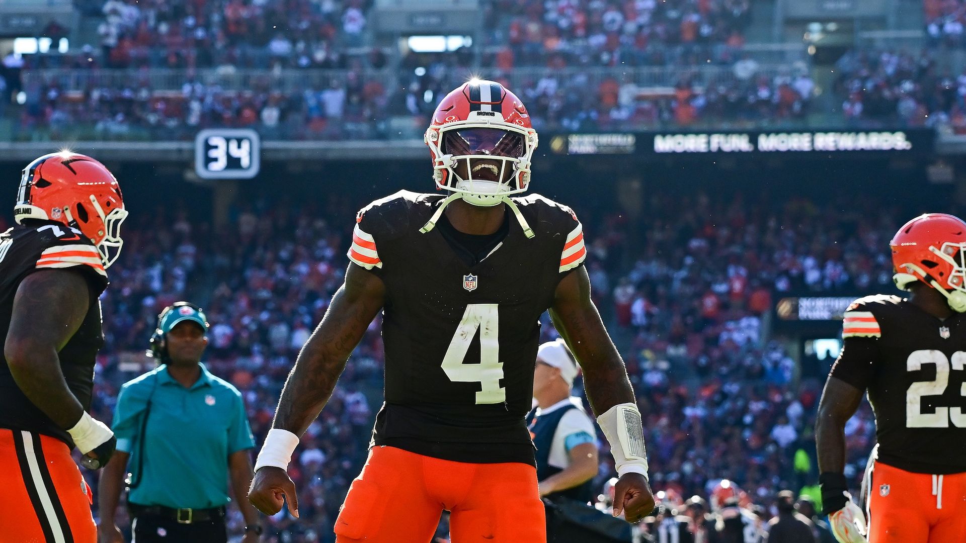 Cleveland Browns players on a sunlit football field, wearing black jerseys and orange pants, centered by a tall player in number 4; crowd fills the stadium stands in the background.