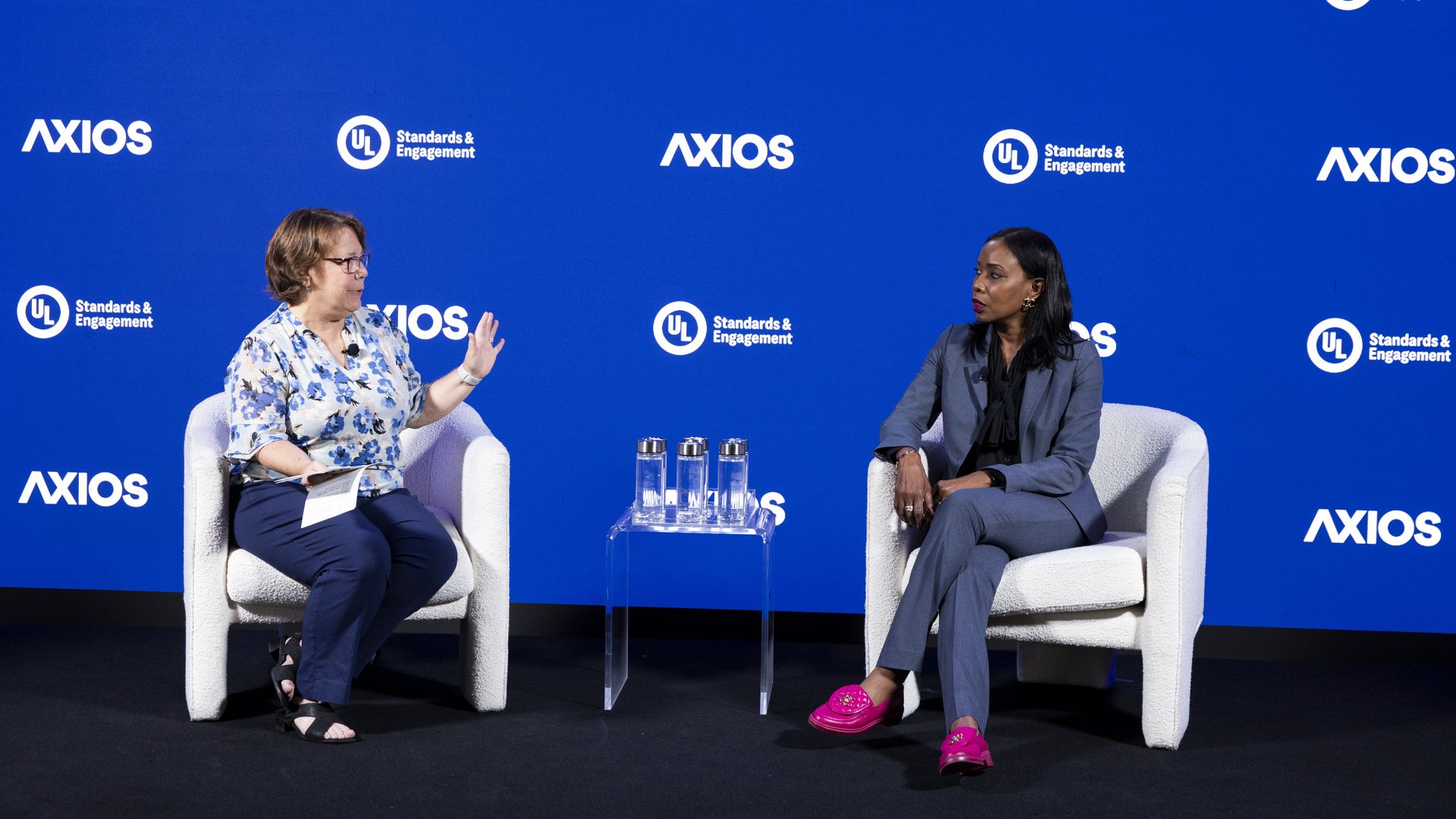 Two women sit in white armchairs on a blue backdrop with AXIOS and UL Standards & Engagement logos; a small glass table with four water bottles sits between them as they chat.
