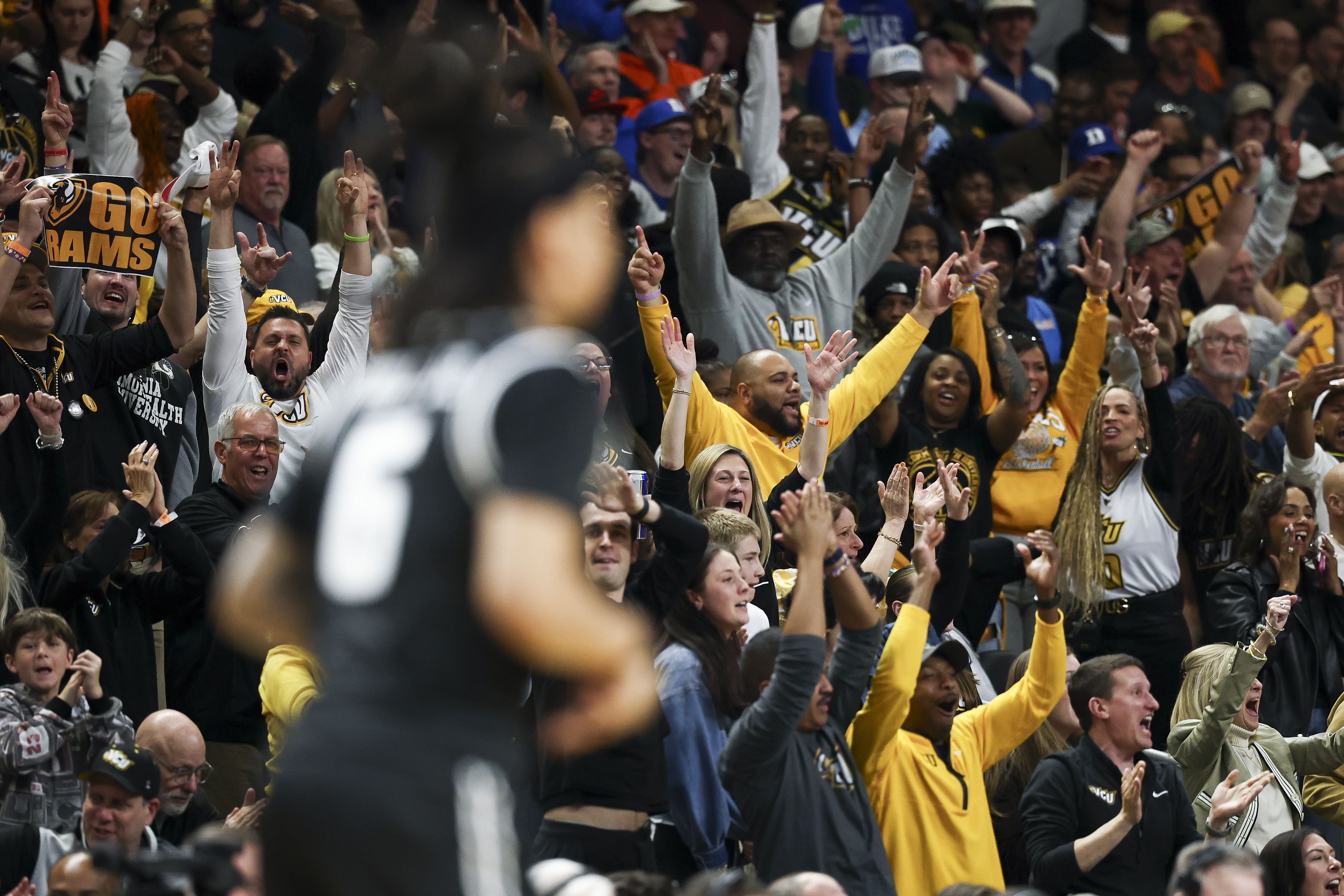 Crowded sports arena filled with cheering fans wearing black and gold, many with hands raised in celebration. A "GO RAMS" sign is visible; foreground figure is blurred as the crowd cheers.