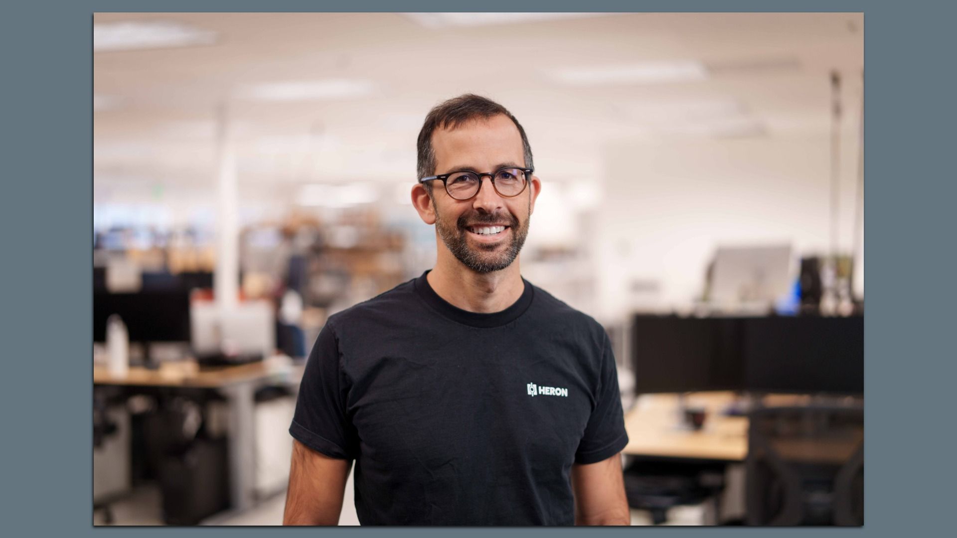 Smiling man with glasses and a short beard, wearing a black t-shirt with a small "HERON" logo, in a bright office with desks blurred in the background.