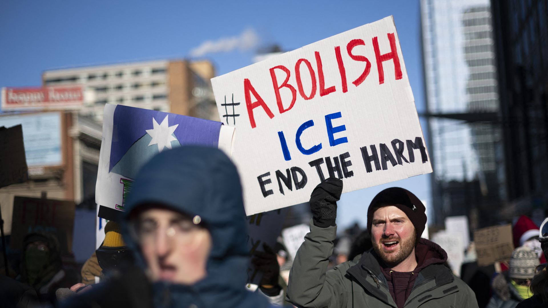 Protesters holding signs outside, with one person holding a sign that reads "#Abolish ICE End The Harm" 