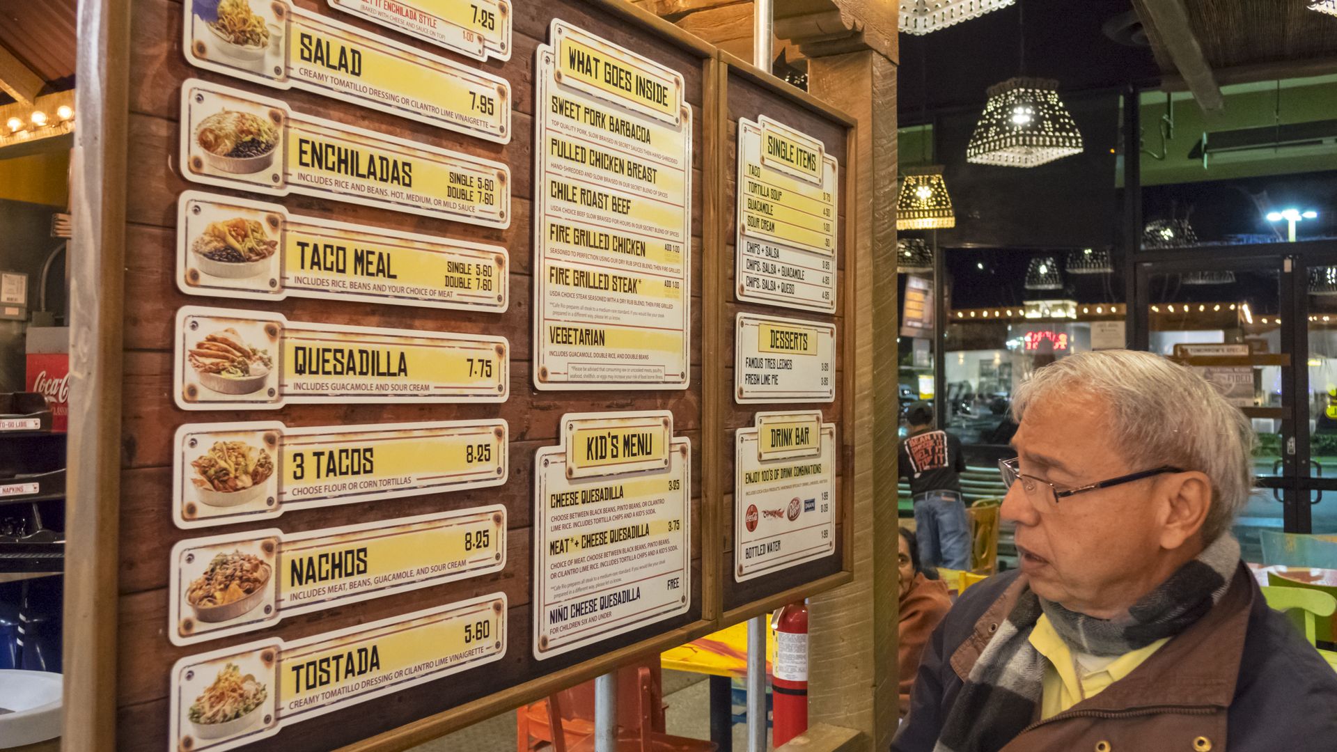 Older man with glasses stands beside a wooden menu wall covered with bright yellow signs listing burrito, salad, enchiladas, tacos, quesadillas; warm overhead lights in a casual restaurant.