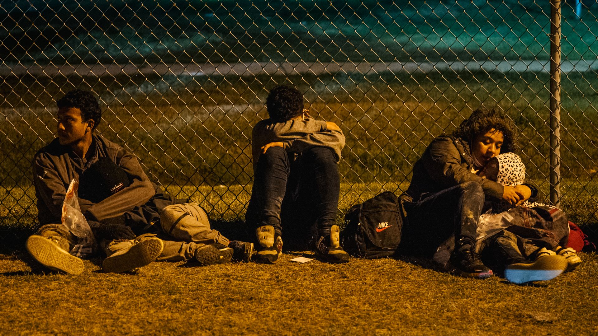 Migrants sit against a fence while waiting to board a border patrol bus after crossing the Rio Grande into the U.S.