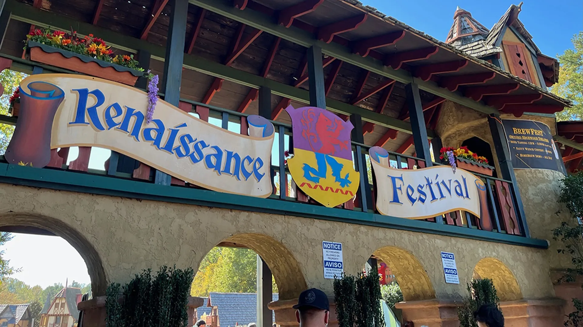Arched entrance to a Renaissance Festival with a banner in blue text reading "Renaissance Festival" and a colorful crest in the center, flowers in window boxes, and a clear blue sky.