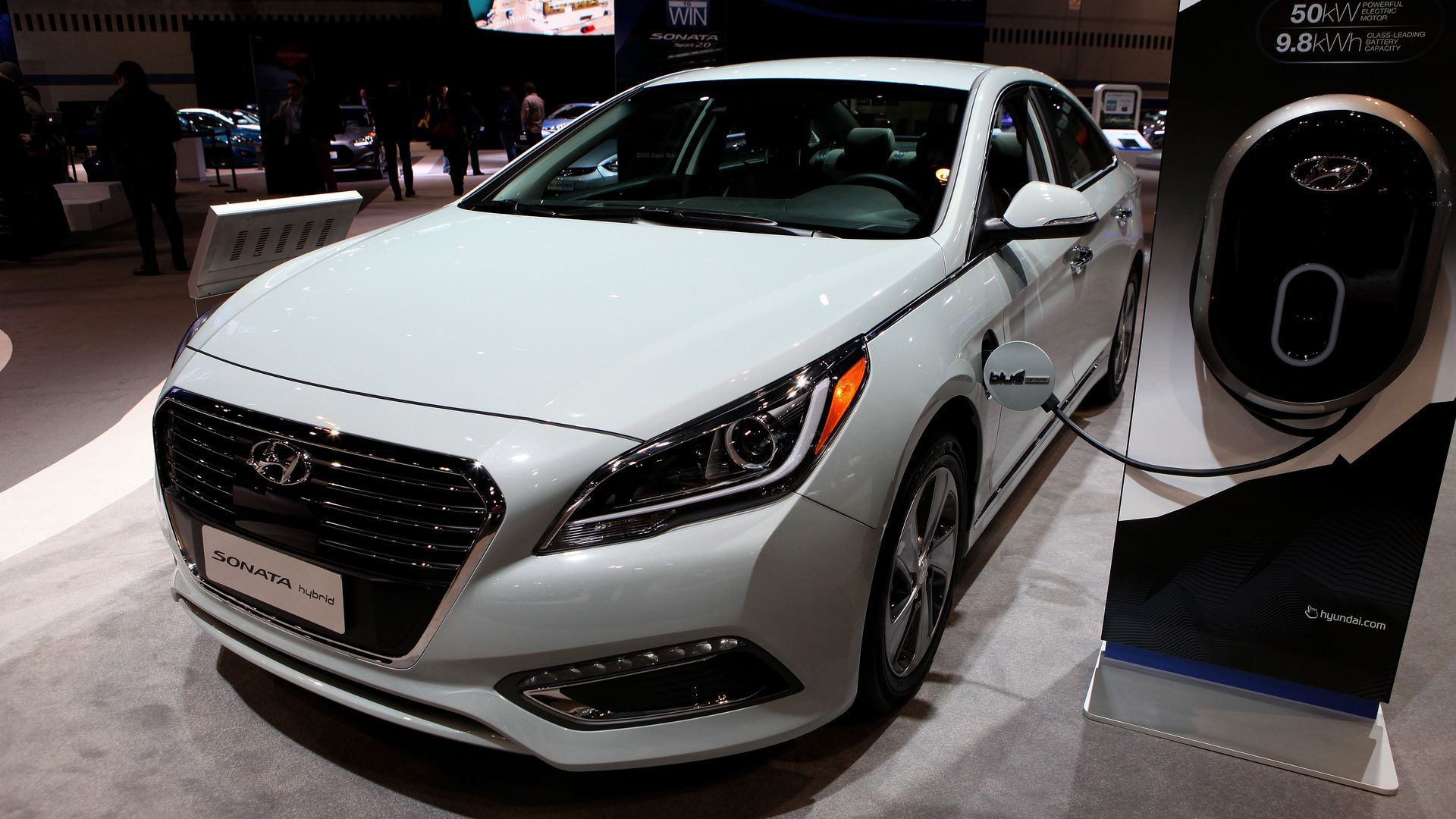 A white vehicle sits on the floor of an auto show with a plug plugged into a charging device