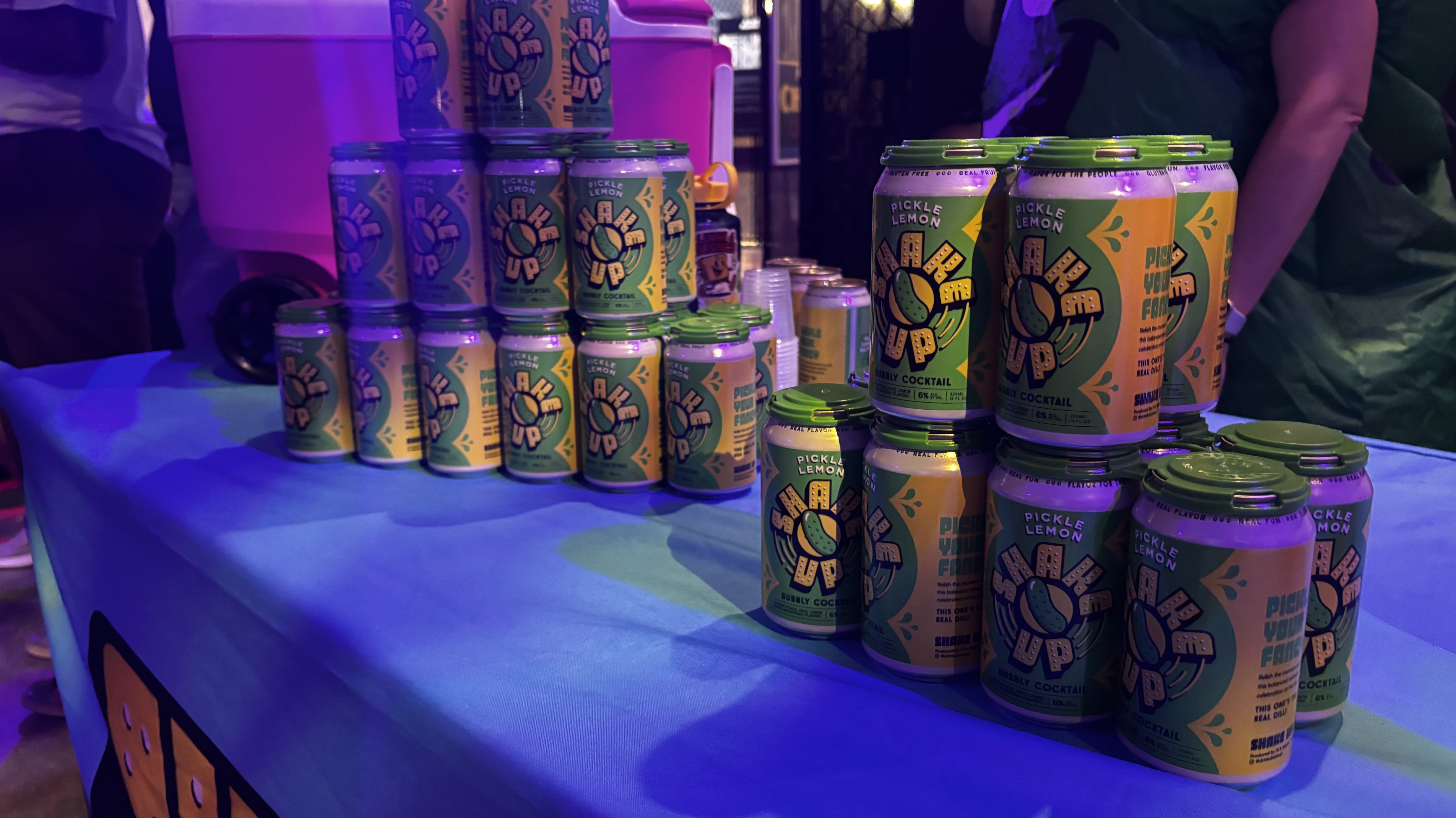Stack of green and yellow cans labeled "PICKLE LEMON" arranged on a blue-lit table at a booth, with purple lighting and blurred people in the background.