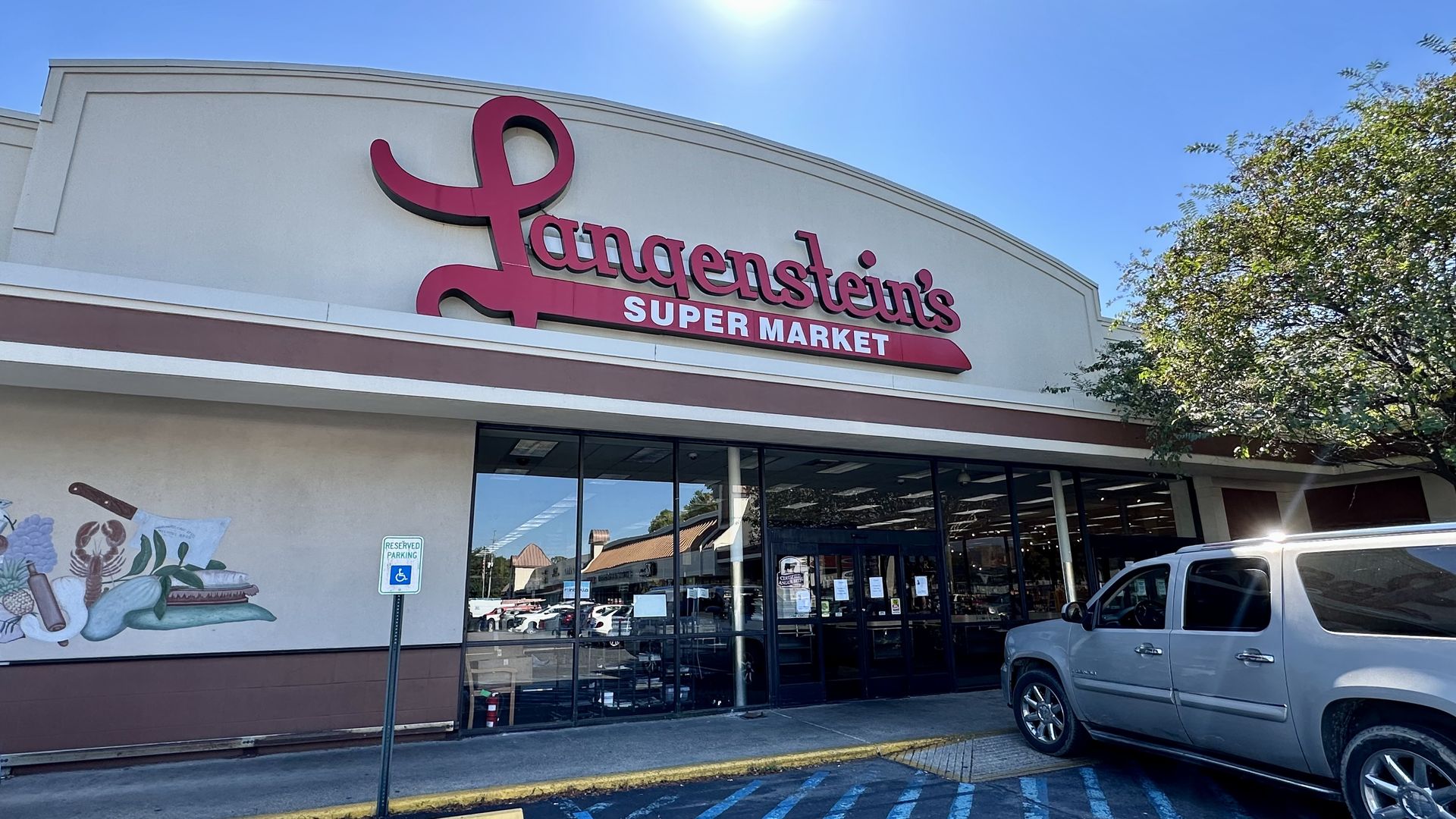 Exterior of Langenstein's Super Market with a red and white sign, clear blue sky, a tree on the right, and a silver SUV parked in a handicapped parking space.