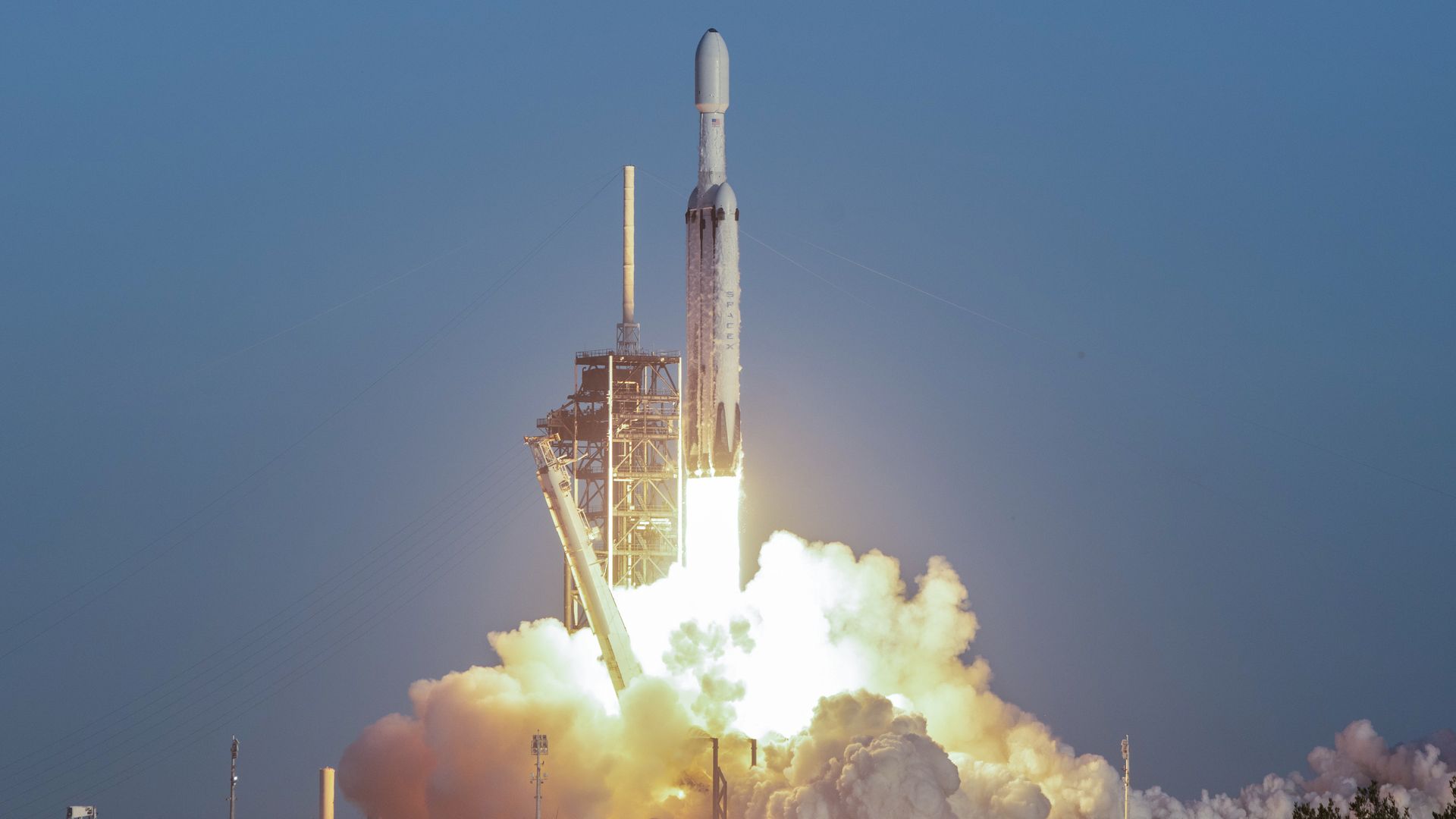 A tall white rocket lifts off from a launch pad, surrounded by bright flames and billowing white smoke, against a clear blue sky.