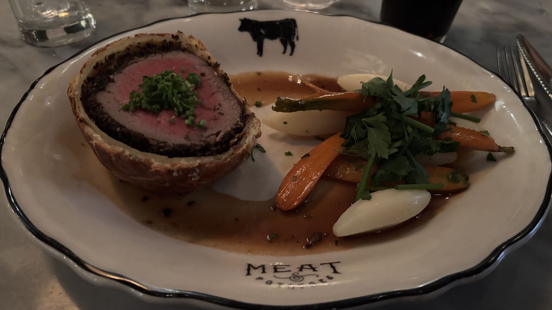 Beef Wellington slice with a golden puff pastry crust and a rosy pink center, topped with chopped chives. Served with glazed carrots, onion wedges, parsley, and a brown jus on a white plate.