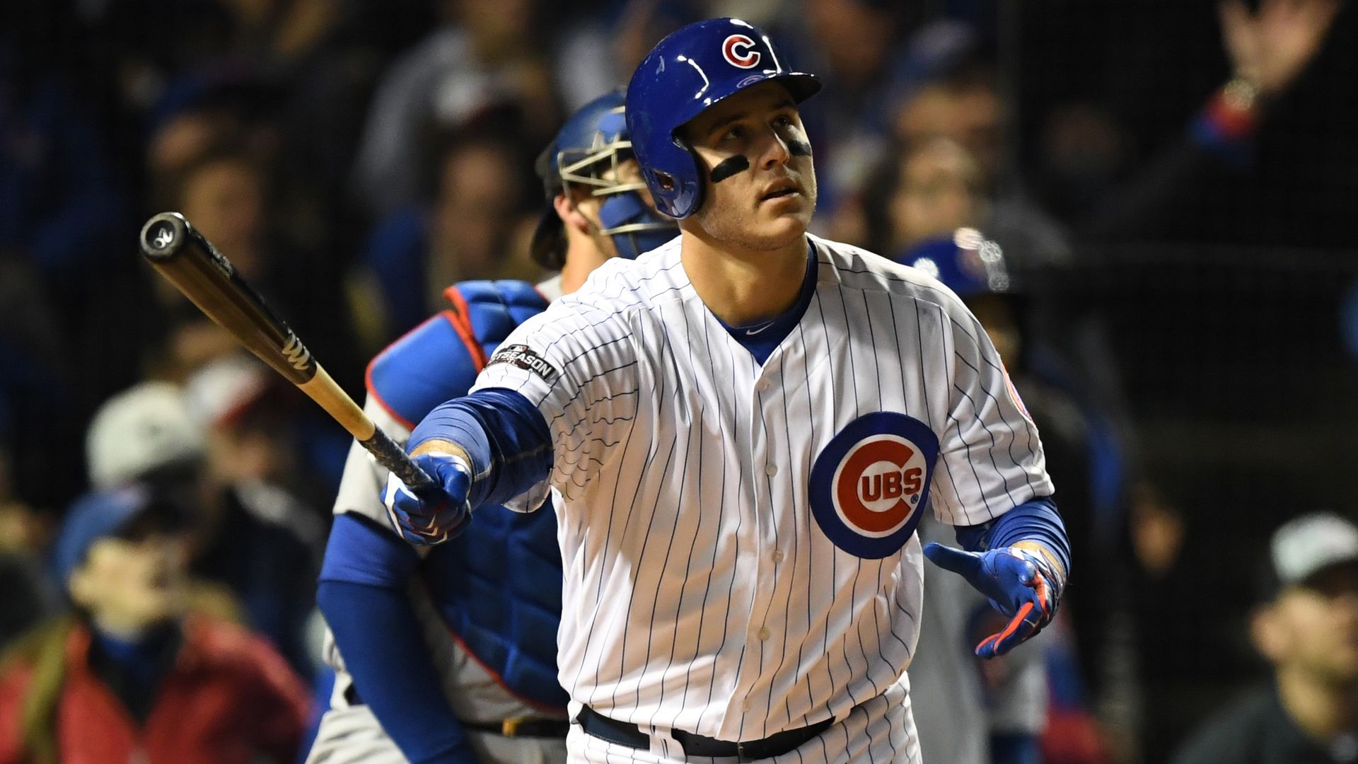 Chicago Cubs baseball player in white pinstripe uniform and blue helmet watching the ball after a swing, with a catcher in blue gear behind him and blurred crowd in the background