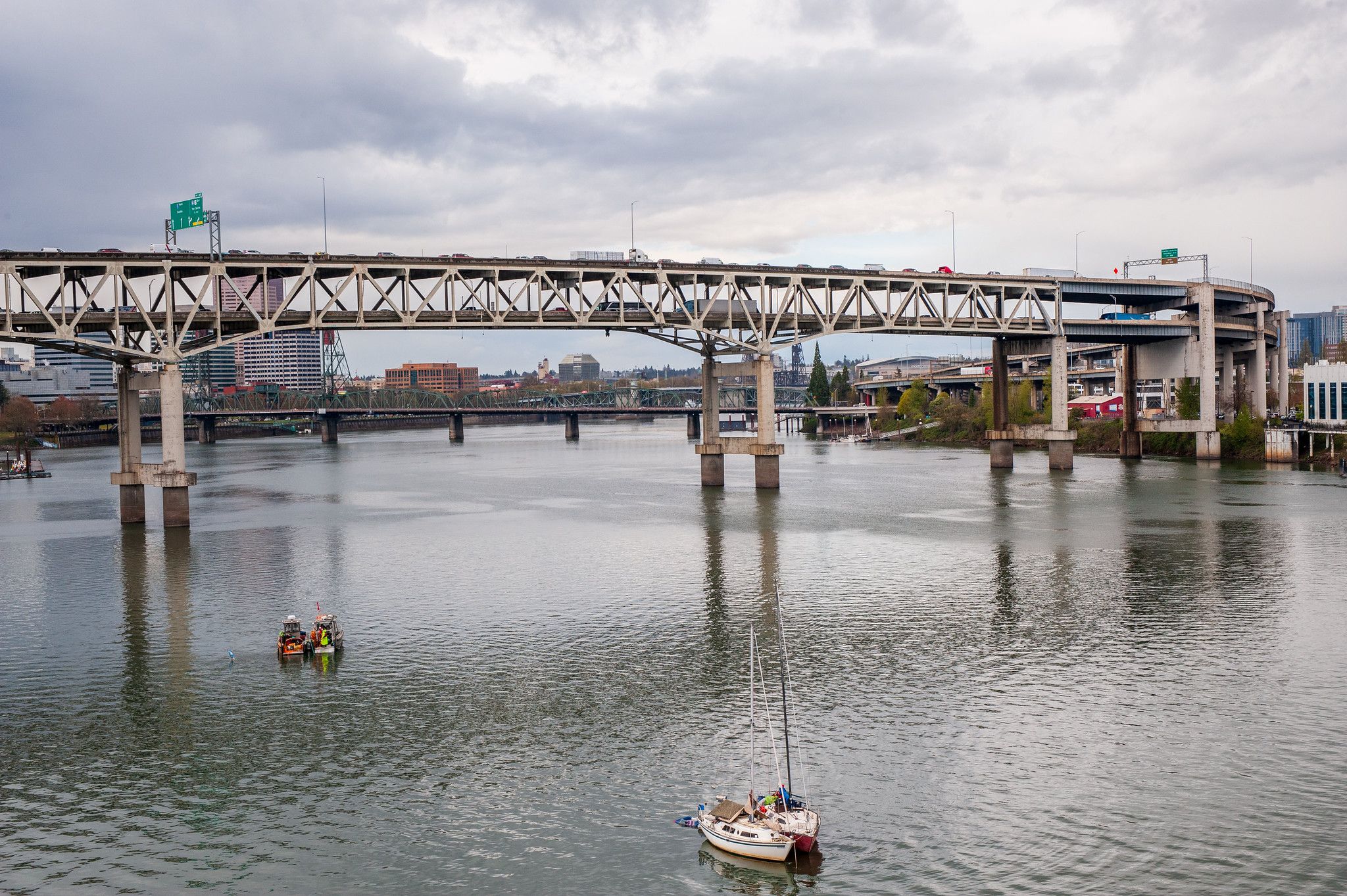 Wide river under a large concrete, multi-level highway bridge; cars travel along the top. Overcast sky, distant city buildings, and small boats on the calm water.