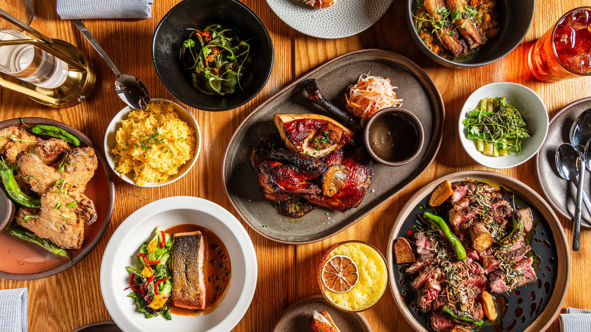 Table with an assortment of dishes including grilled chicken, fried chicken wings, yellow rice, seared salmon, steak with potatoes, asparagus, small appetizers, a drink with ice, and various garnishes on wooden surface.