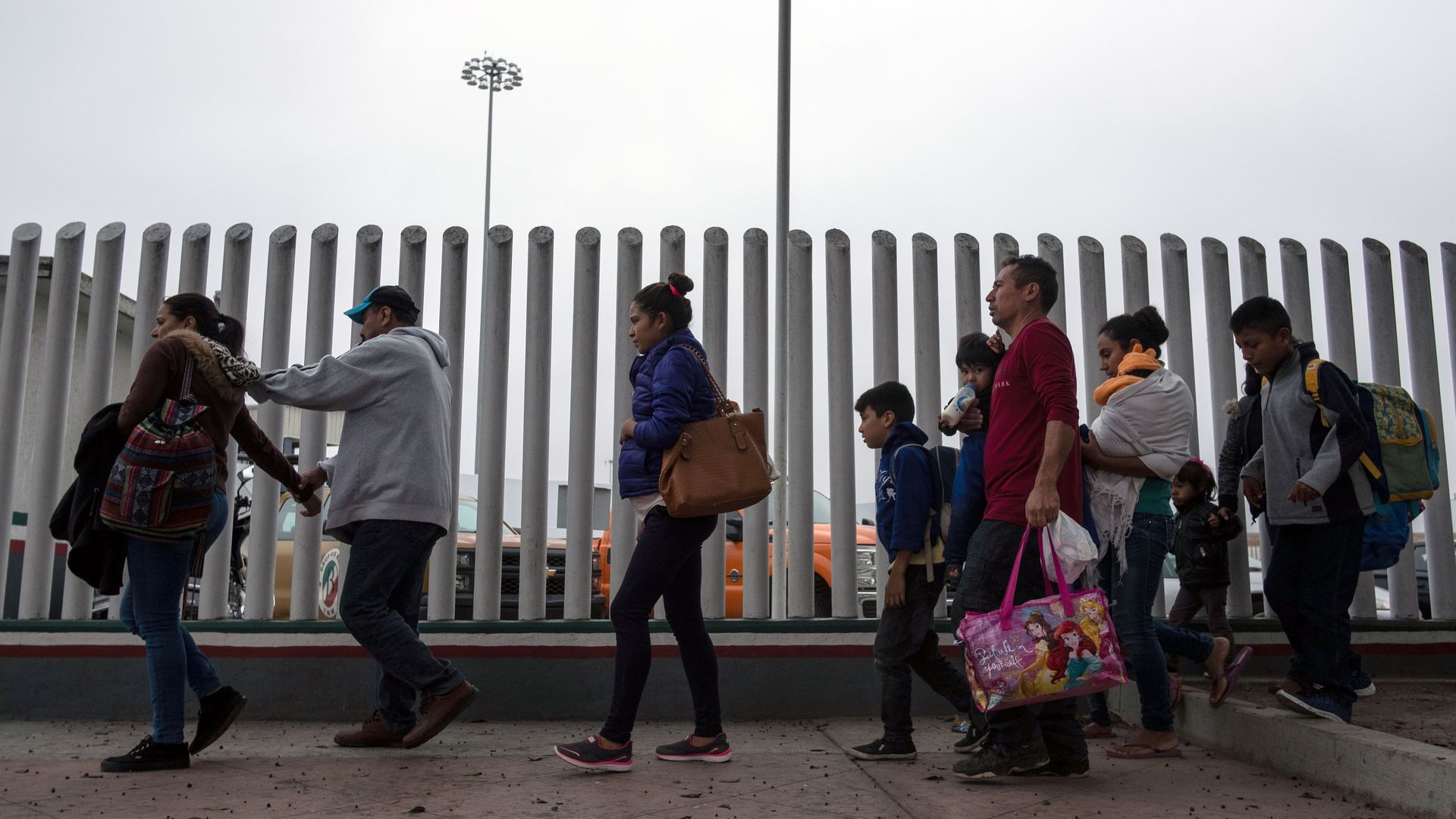 A line of adults and children walk toward a port of entry with a fence next to them. 