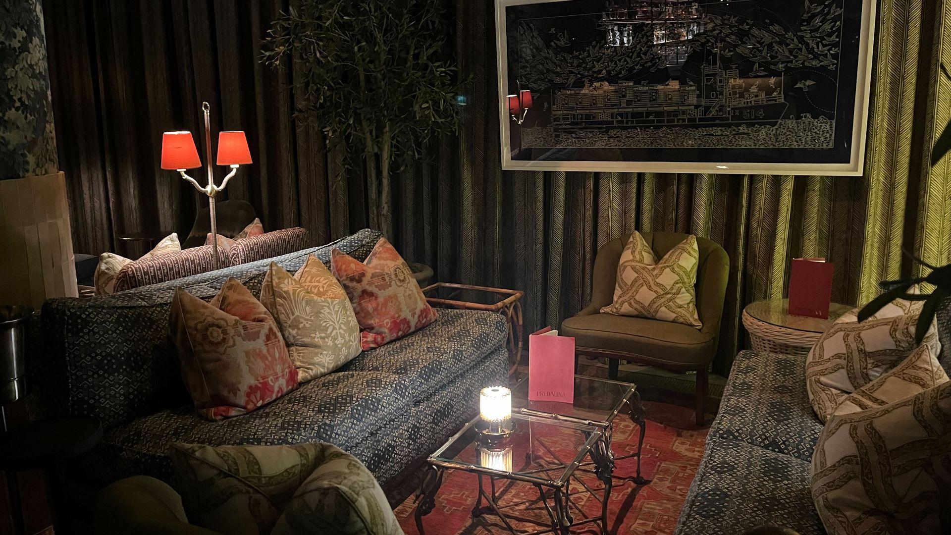 Dimly lit cozy lounge with patterned sofas, floral and chain-patterned pillows, small glass tables, red lampshades, a large framed ship artwork, and a red area rug.