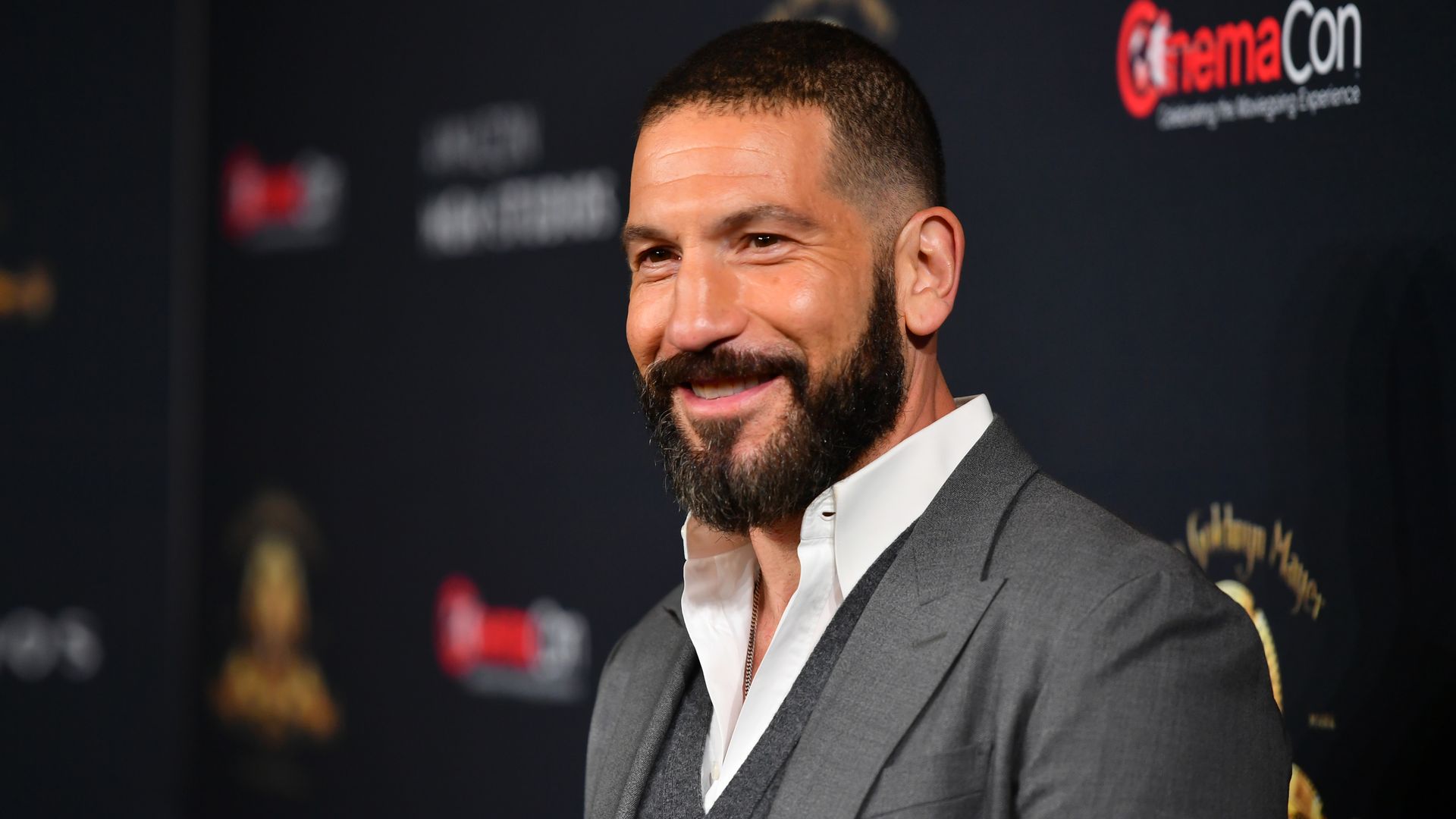 Jon Bernthal smiling with short dark hair and beard wearing a gray suit, white shirt, and gray vest, posing at a CinemaCon event with a dark background featuring logos.