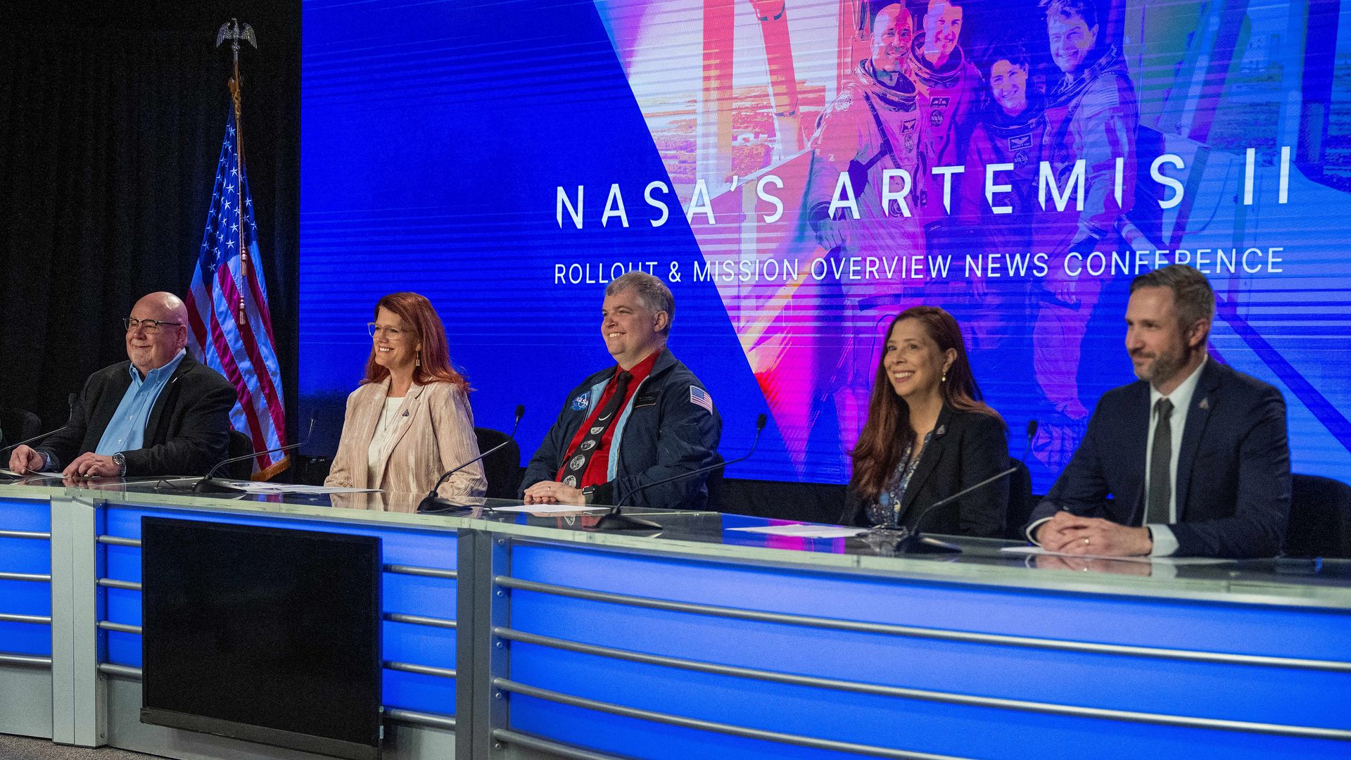Five NASA employees sit at a blue curved desk while a large screen behind displays the title "NASA's Artemis II Rollout & Mission Overview News Conference."