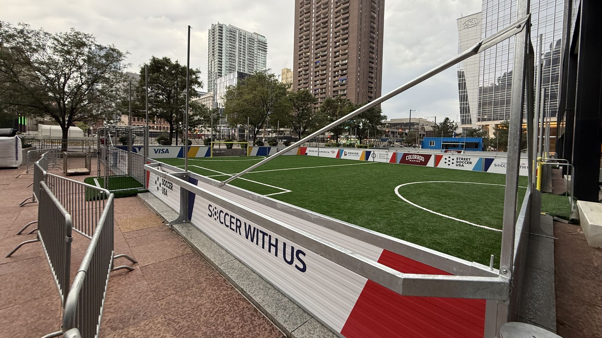 Small outdoor soccer field with green artificial turf surrounded by metal railings and advertising boards. Urban setting with tall buildings and cloudy sky in the background.