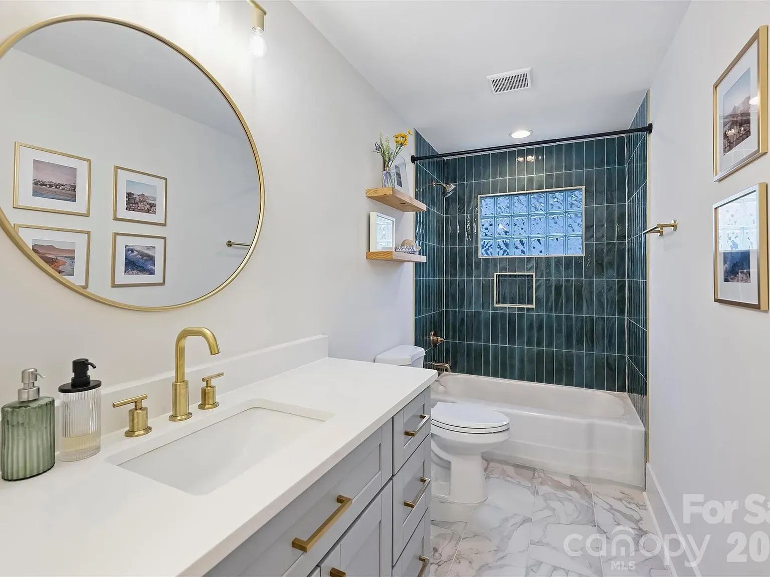 Bright modern bathroom with a large round gold-framed mirror above a white vanity and gold fixtures. Teal-tiled shower with a glass-block window, gold hardware, and marble flooring.