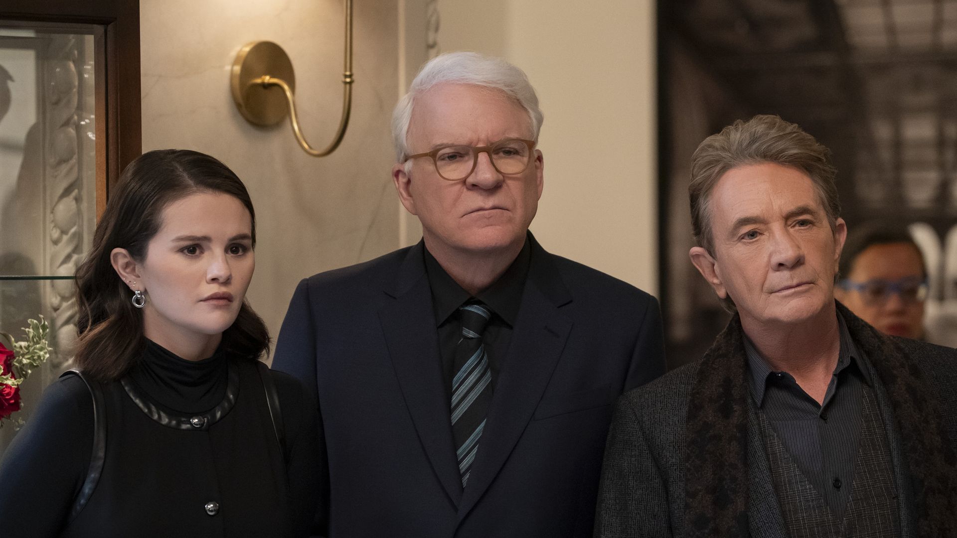 Three people stand side by side indoors, a young woman with dark hair in black, a white-haired man with glasses in a dark suit, and a man with light brown hair in a dark patterned coat, all serious.