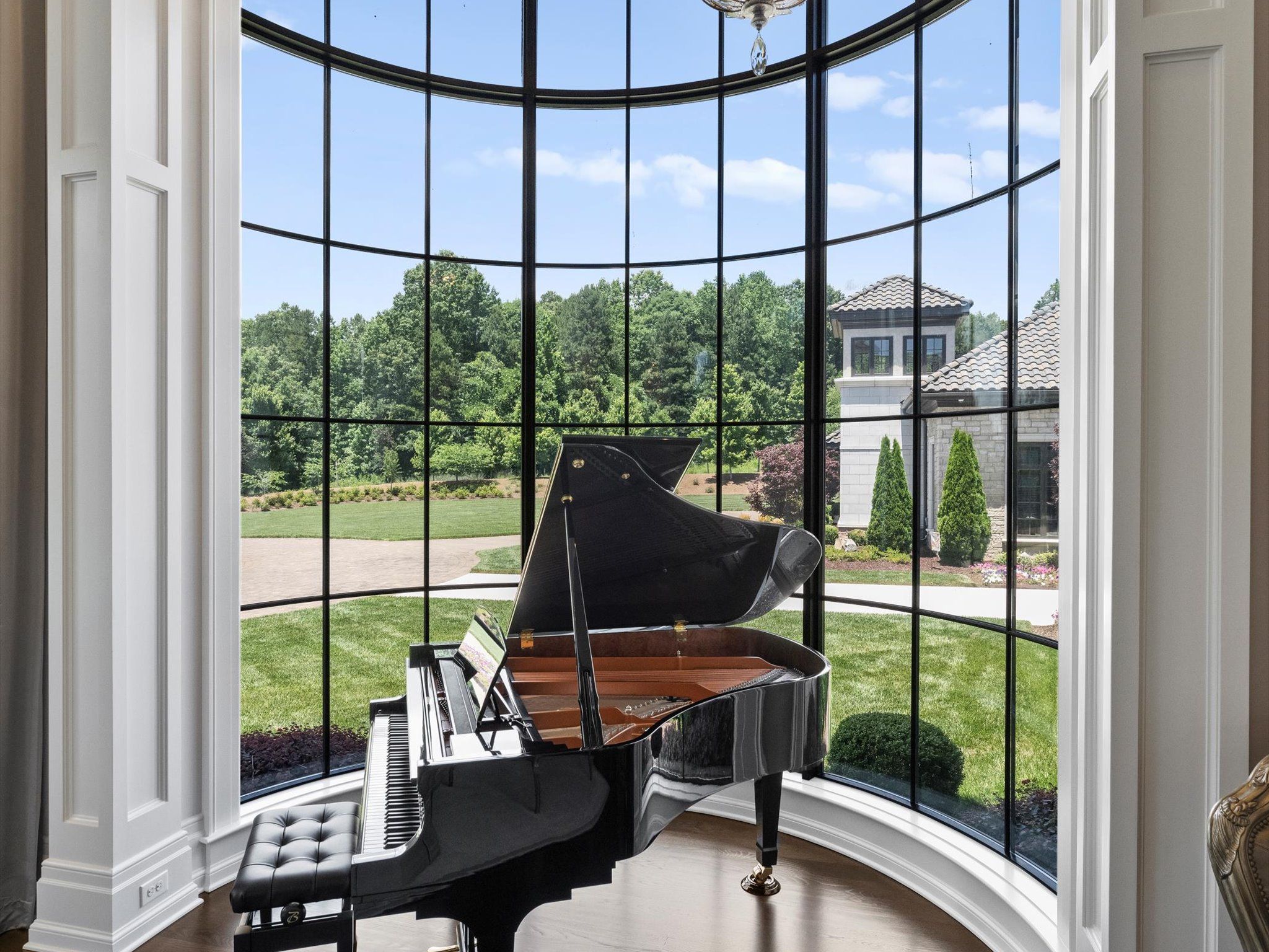 Black grand piano with tufted bench in front of large curved floor-to-ceiling windows showing green lawn, trees, and part of a house with stone walls and tiled roof outside on a sunny day.