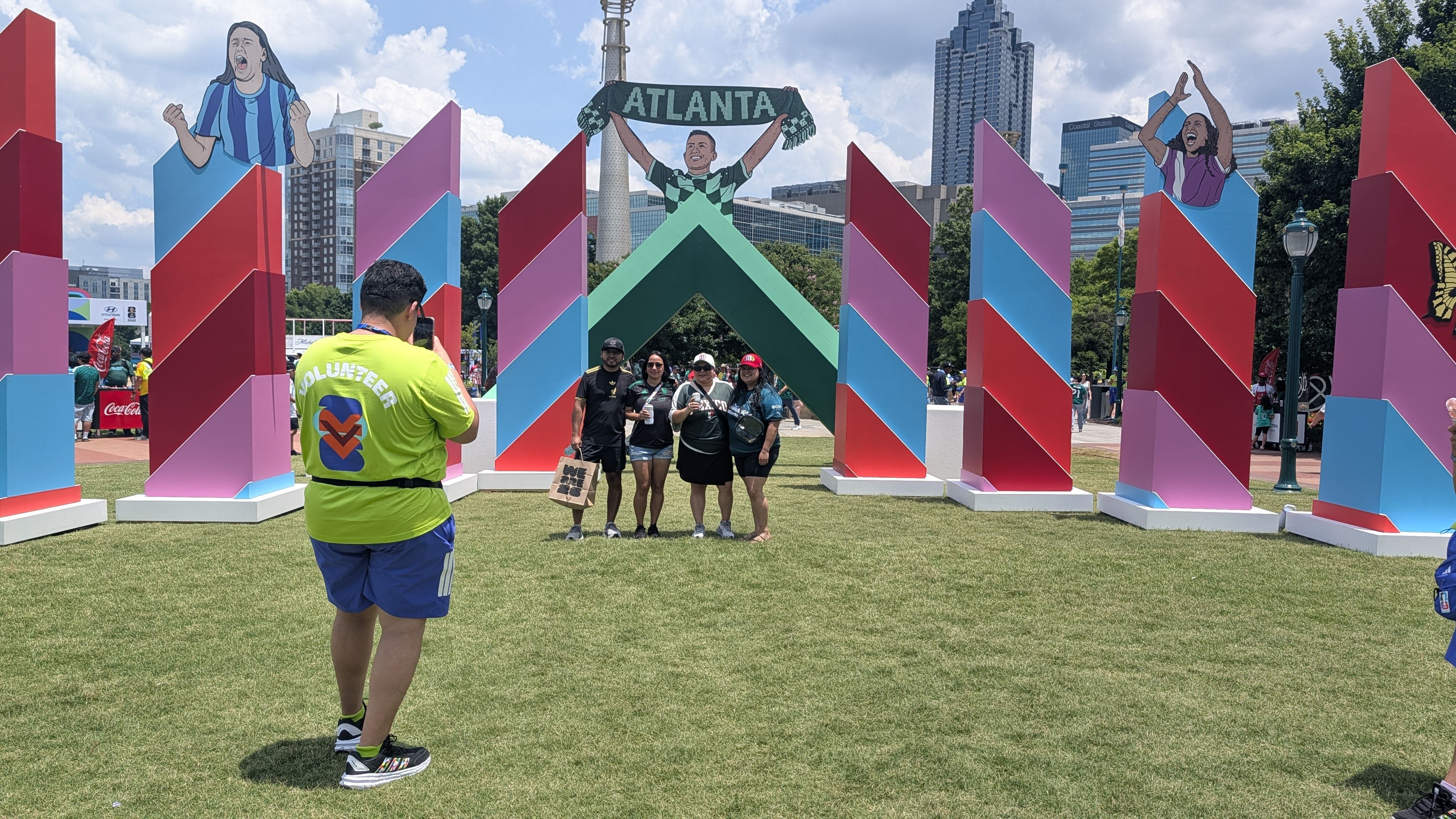 Colorful geometric arch installation in a park. A volunteer in a neon shirt photographs a group posing in front, with a banner reading "ATLANTA" and cartoon figures above a city skyline.