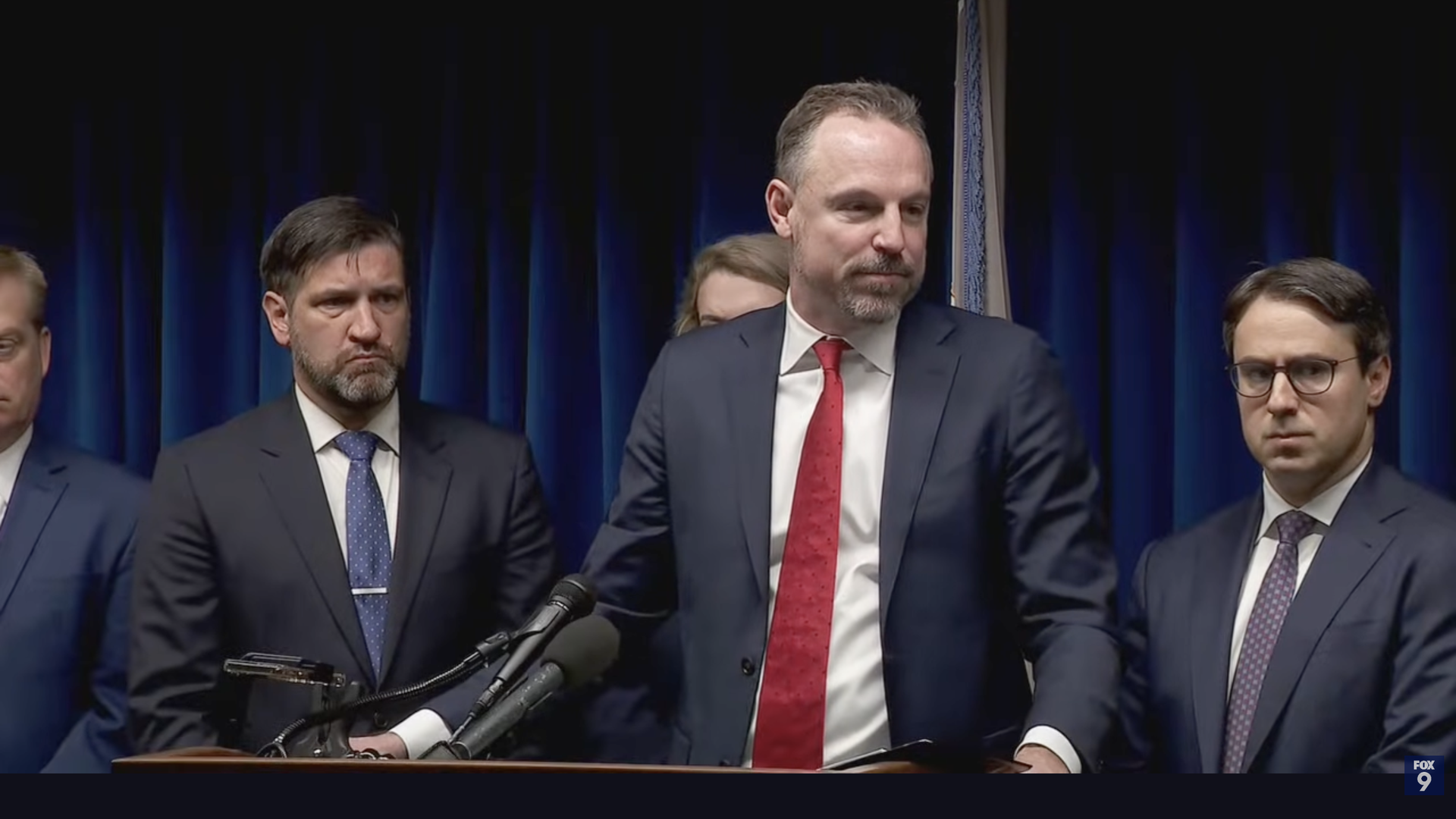 Joe Thompson stands at a podium flanked by two men. He's wearing a dark blue suit and red tie.