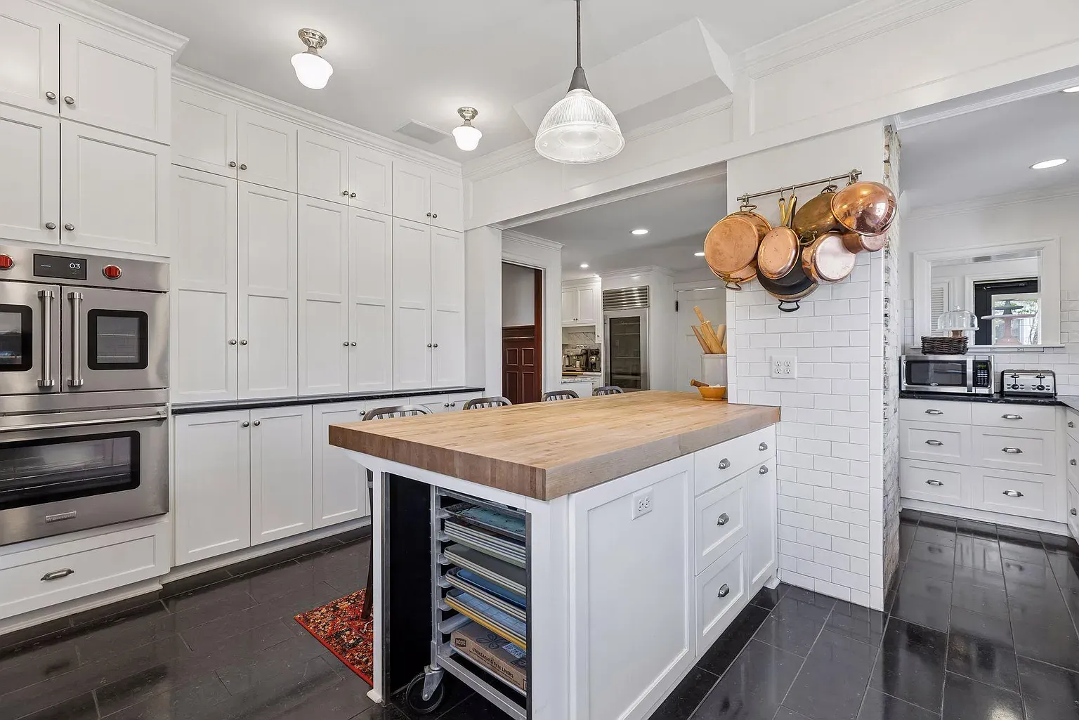Bright white kitchen with tall white cabinets, stainless steel double ovens, and a butcher-block island. Copper pots hang over a white subway tile wall; dark flooring and a pendant light.