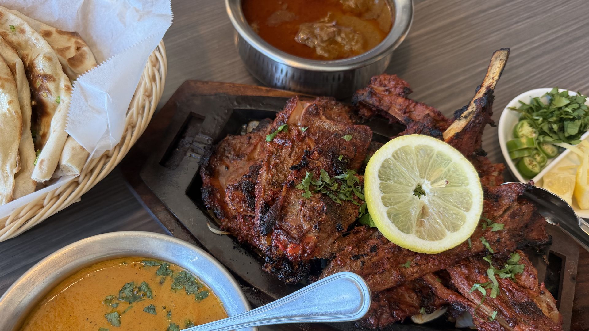 Basket of warm naan bread, charred tandoori chicken slices with a lemon half, a metal bowl of curry, and a small side of chopped cilantro on a dark wooden table.