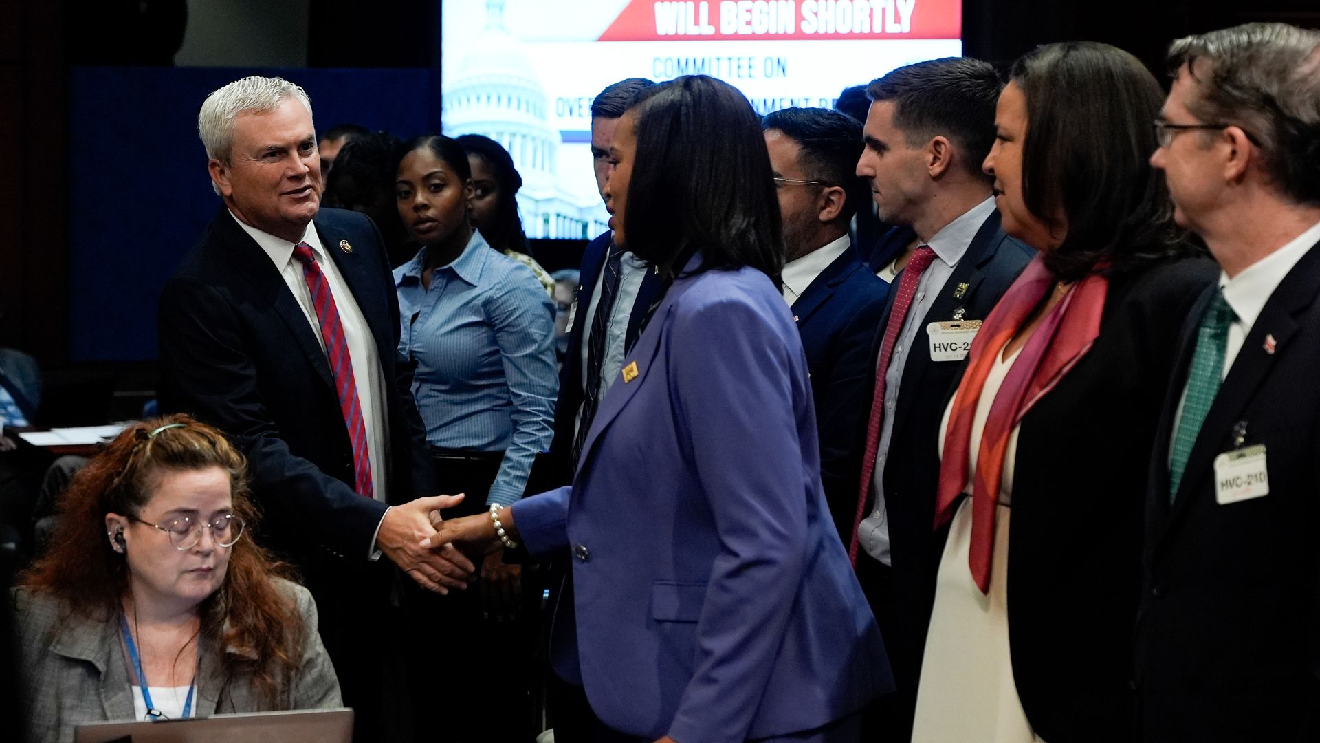 In a hearing room, Rep. James Comer shakes the hand of Mayor Muriel Bowser, surrounded by her aides