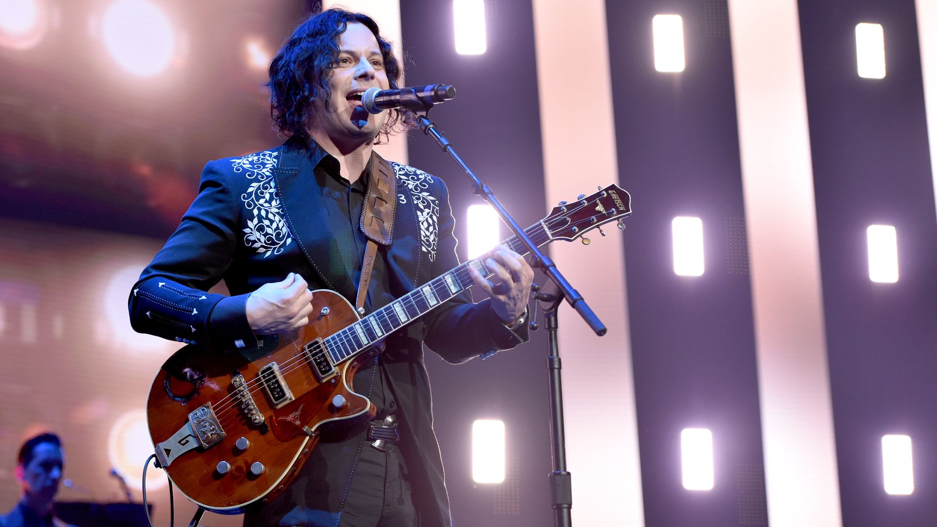 A man with a guitar sings into a microphone nearby.