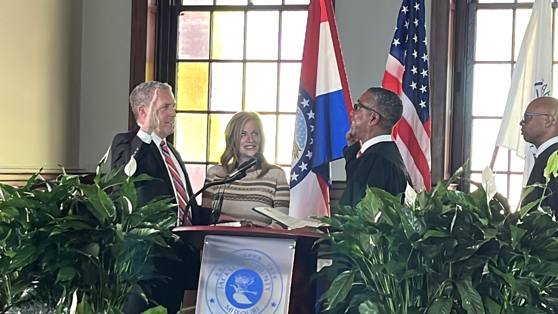 Man in suit raising right hand, woman in striped sweater smiling, man in judge robe with glasses raising right hand, another judge nearby, flags of Missouri and the USA in background.