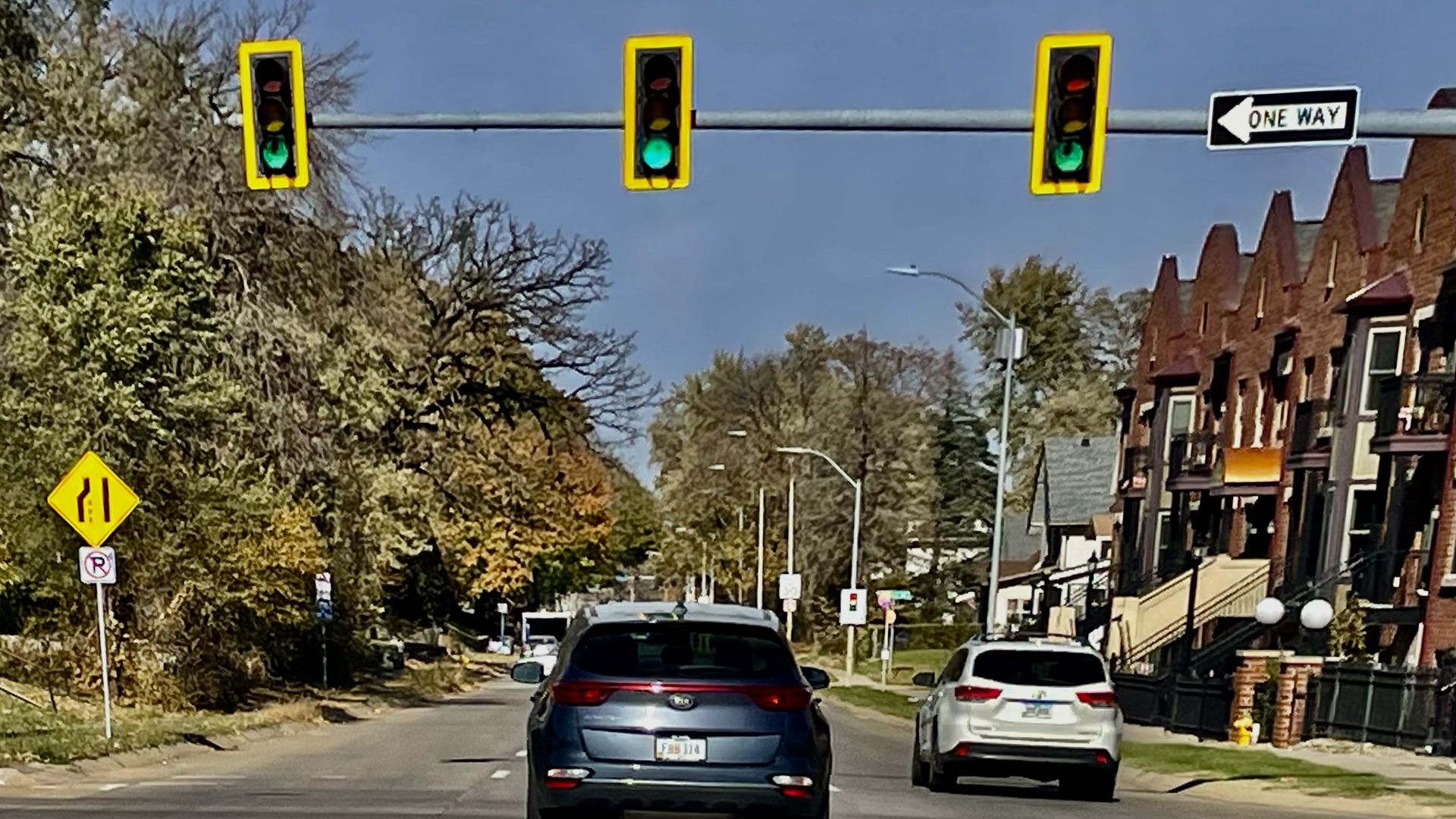Urban street scene with three traffic lights in yellow frames overhead; a one-way sign on the right; red-brick row houses with stairs; two cars on a sunny street, autumn trees on the left.