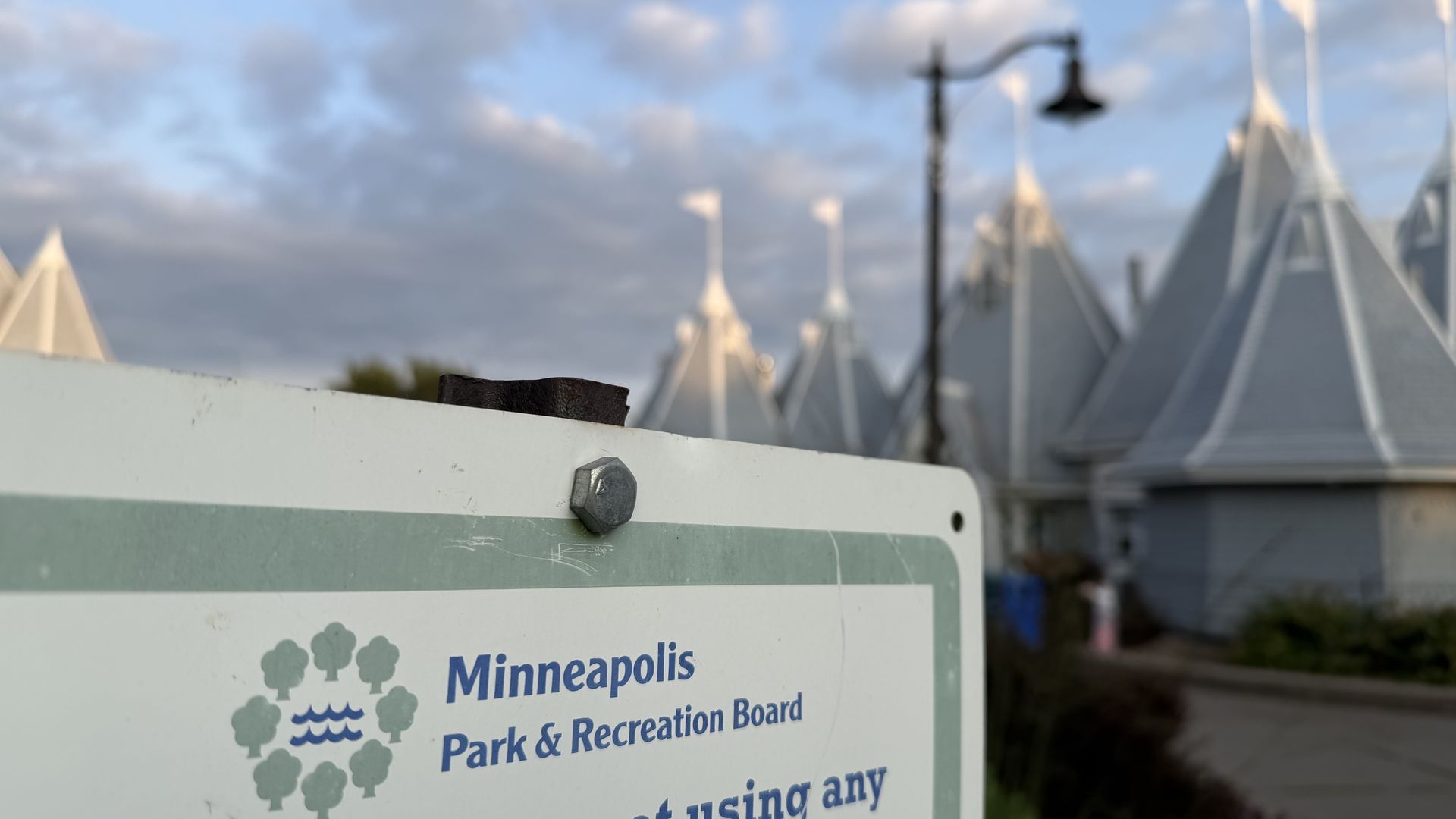 Close-up of a sign that says Minneapolis Park & Recreation Board with a blurred background of white tent shapes, a black street lamp, and a blue sky with clouds.