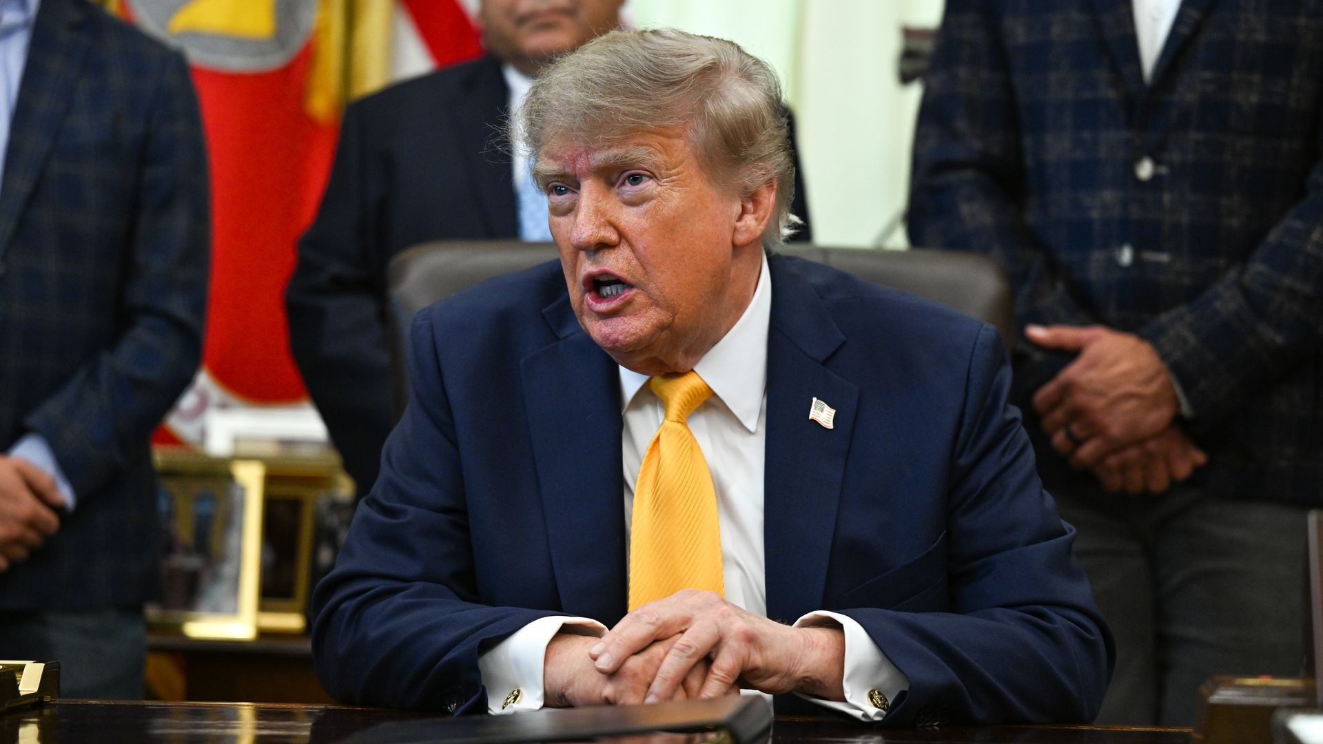 President Donald Trump sits at a desk in a navy suit and orange tie, hands clasped as he speaks, with officials and flags visible behind him.