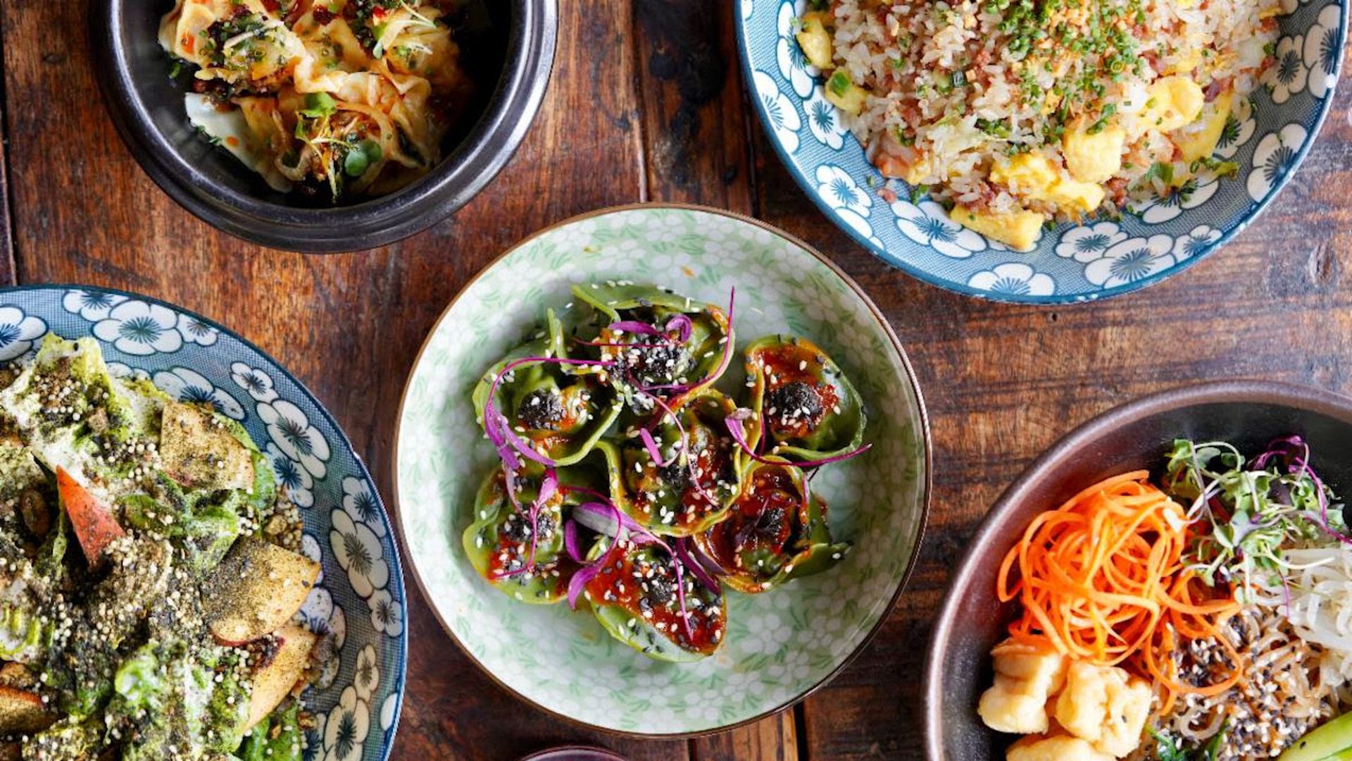 Top-down view of various colorful dishes on a wooden table including green dumplings with sauce and sesame seeds, fried rice, salad with dressing, carrot noodles, and mixed vegetables.