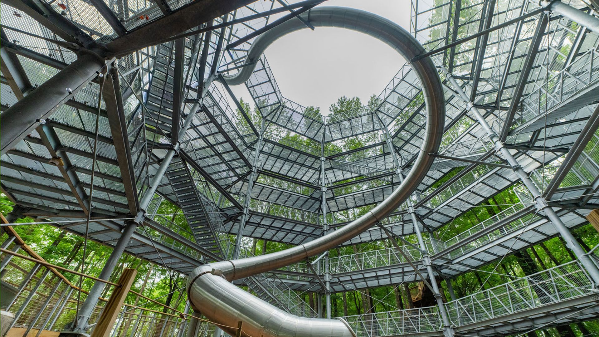 A large, multi-level metal treehouse structure with stairs and walkways surrounds a tall metal slide twisting from the top to the forest floor covered in brown leaves.