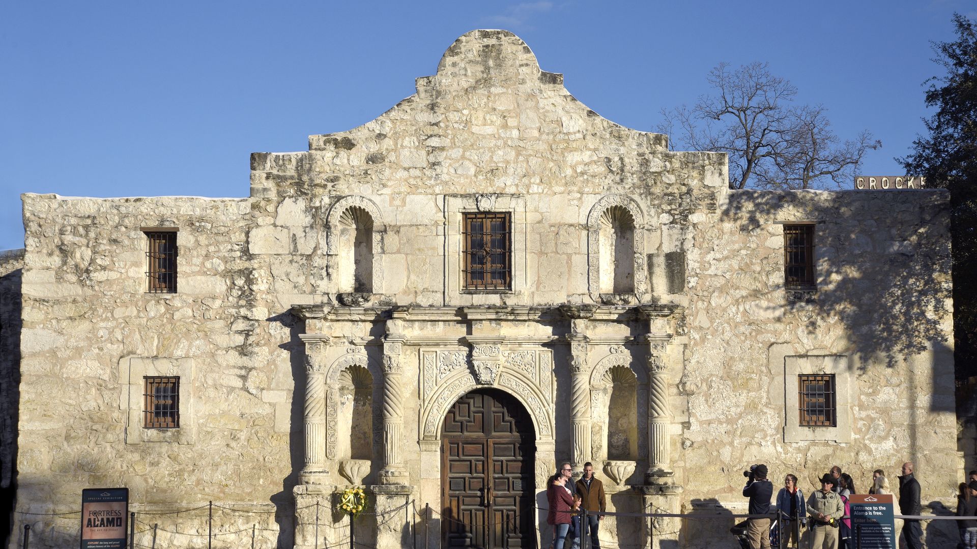 An exterior view of the Alamo with a line of people outside. 