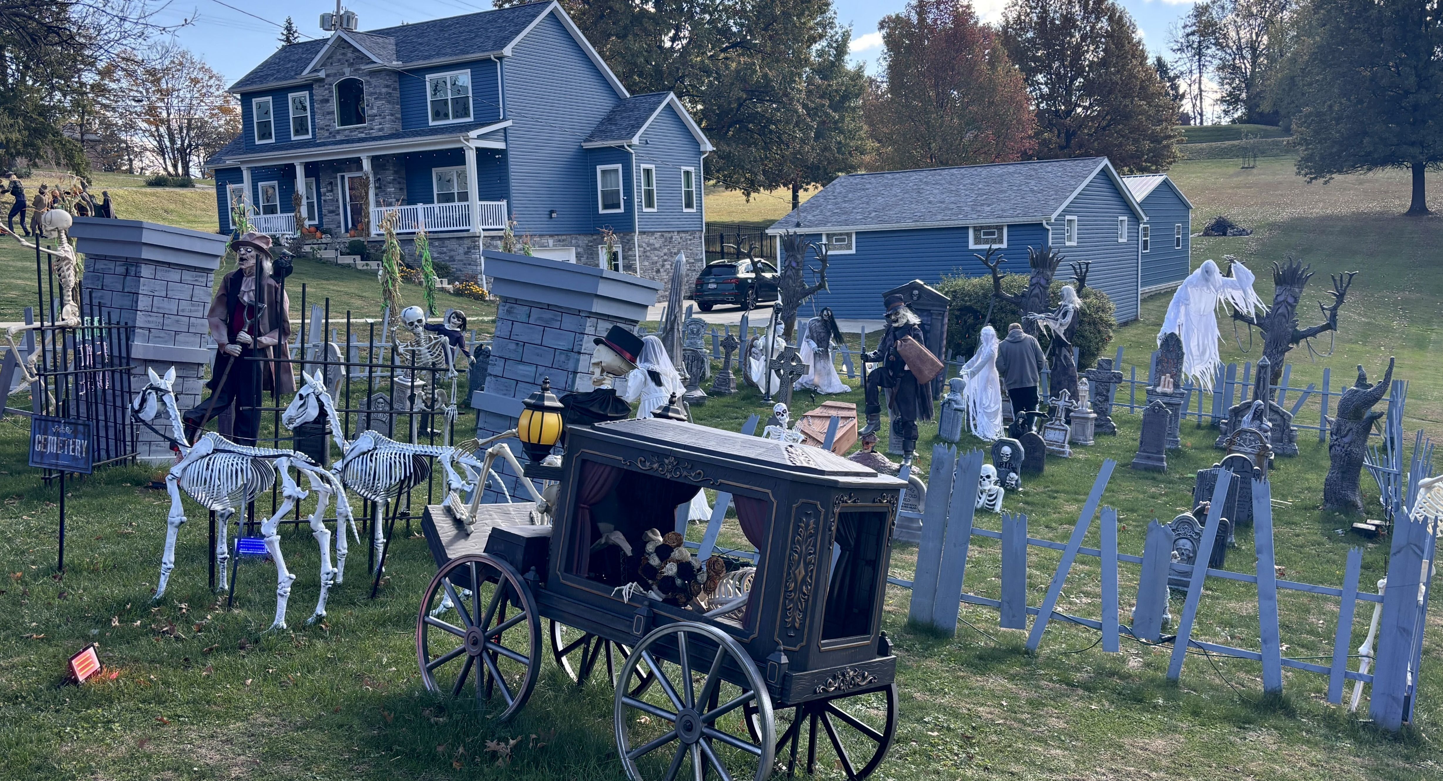 Yard decorated with Halloween cemetery scene including skeletal horses pulling a black hearse, skeletons, ghosts, tombstones, bare trees, and a haunted blue house in the background.