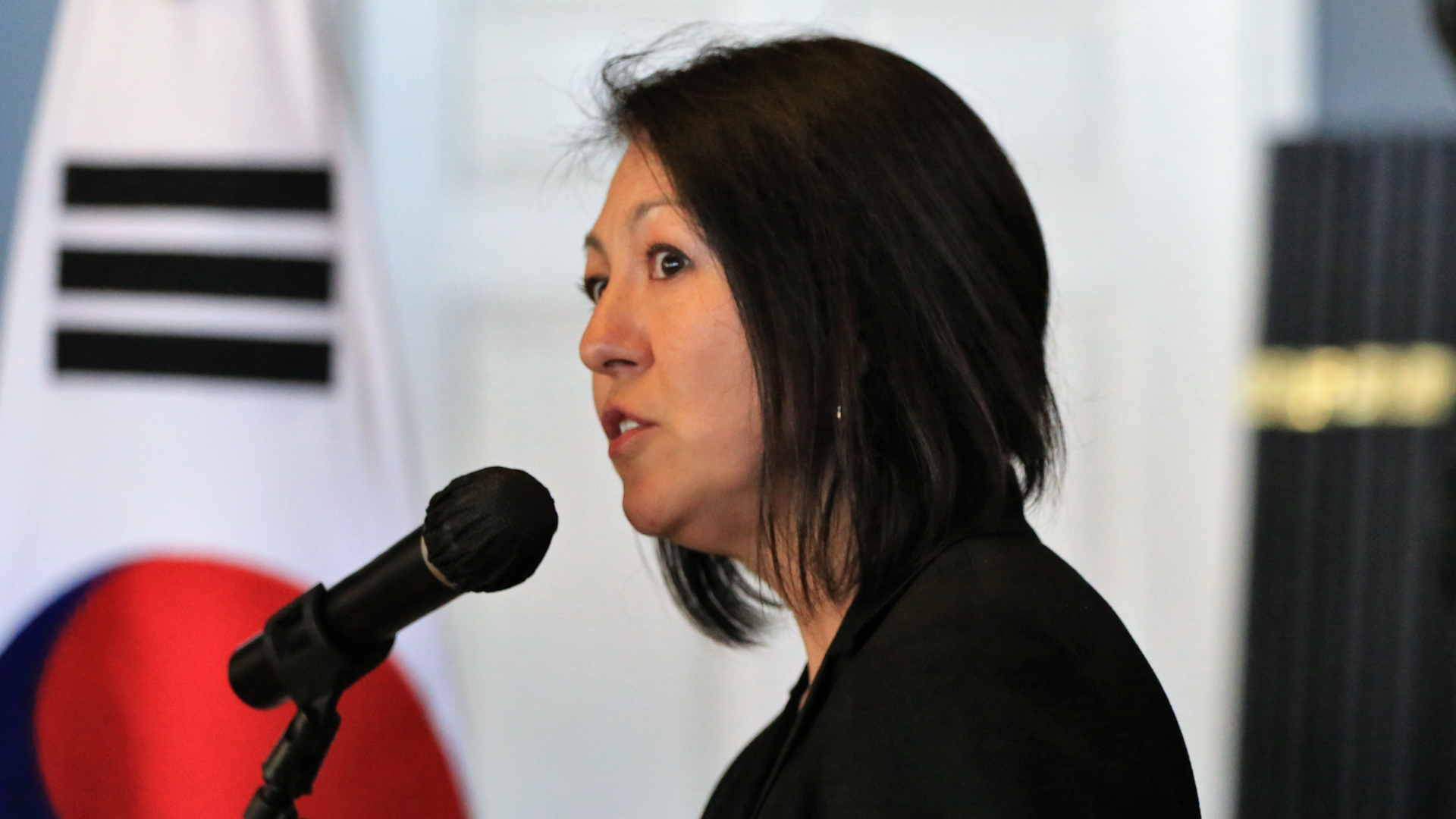 A woman with chin-length dark hair speaks into a microphone, with a Korean flag in background. 