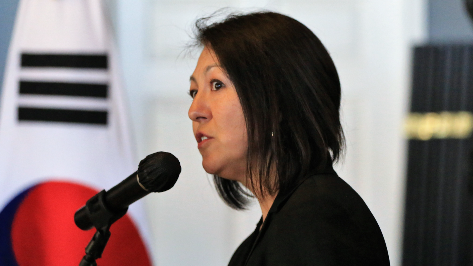 A woman with chin-length dark hair speaks into a microphone, with a Korean flag in background.