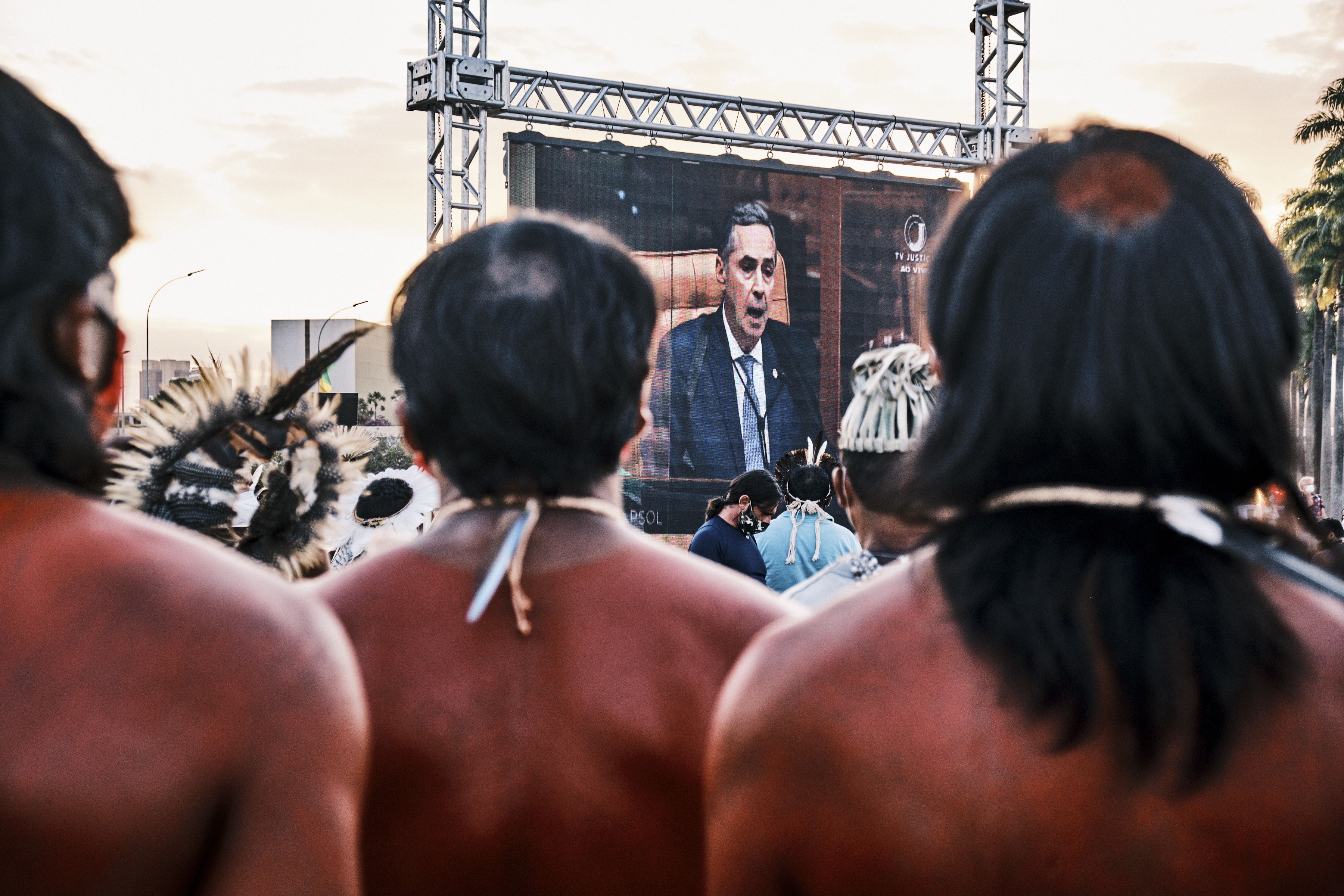 Photo of three Indigenous people watching a large screen showing Supreme Court Justice Luis Roberto Barroso