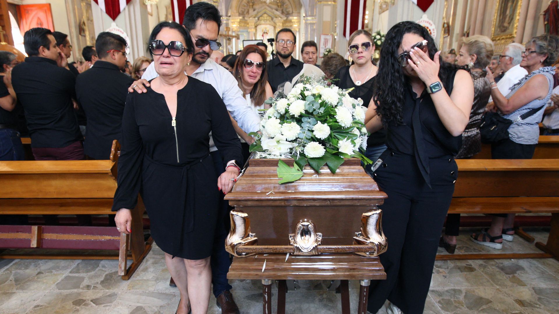 A group of people led by two women who are crying push a brown casket with white flowers atop 