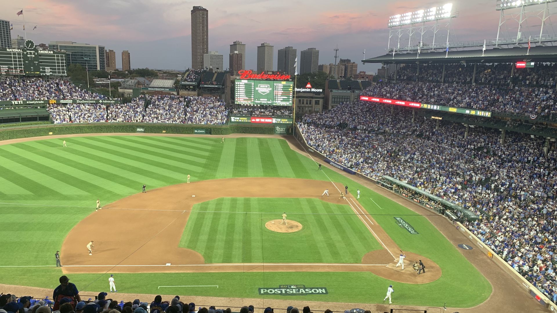 Evening baseball game at a packed stadium with green field, players in position, and city skyline under a colorful sky. Signs for Budweiser and Benjamin Moore visible.