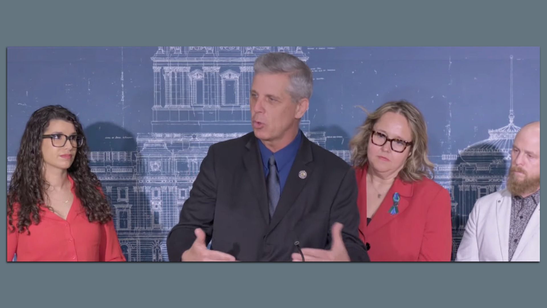 Four panelists at a press briefing in front of a blue architectural backdrop; central man in a dark suit speaks, flanked by two women in red and a bearded man in a light jacket.