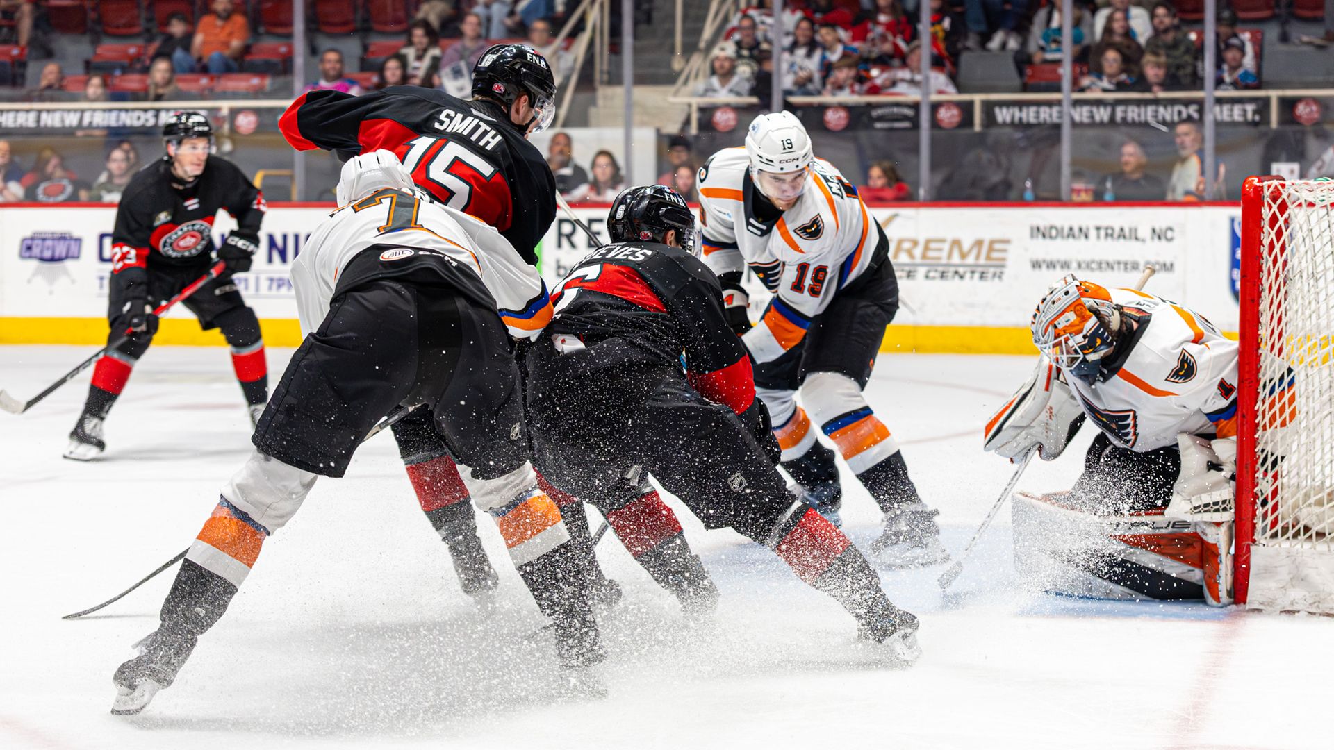 Hockey game in progress: players from opposing teams clash in front of the net while the white-and-orange goalie protects the crease; crowd watches as ice splashes from skates.