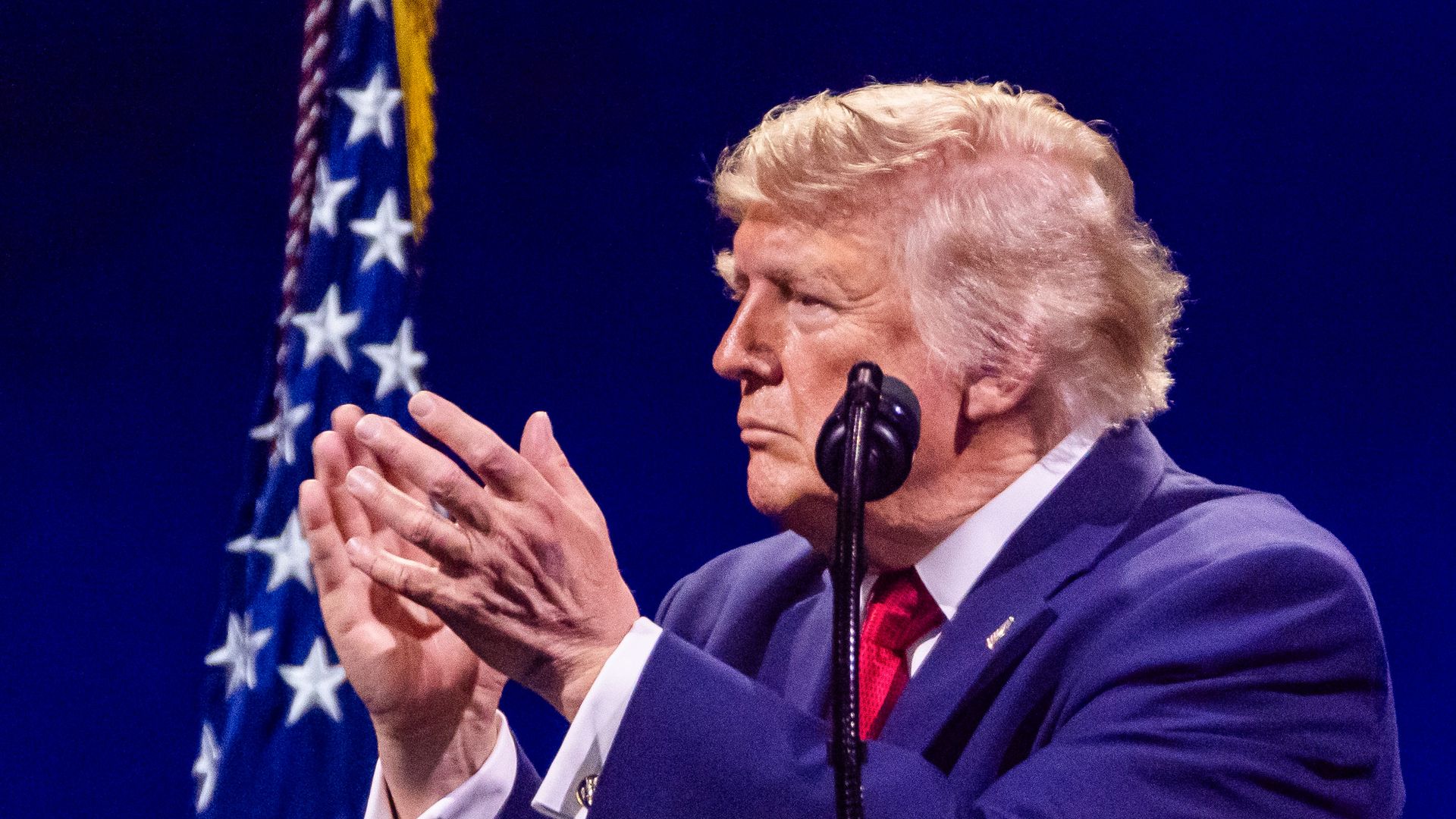 President Trump claps his hands together while wearing a dark blue suit, a white collared shirt, a pin and a red tie and standing behind a microphone and in front of an American flag on a stage.