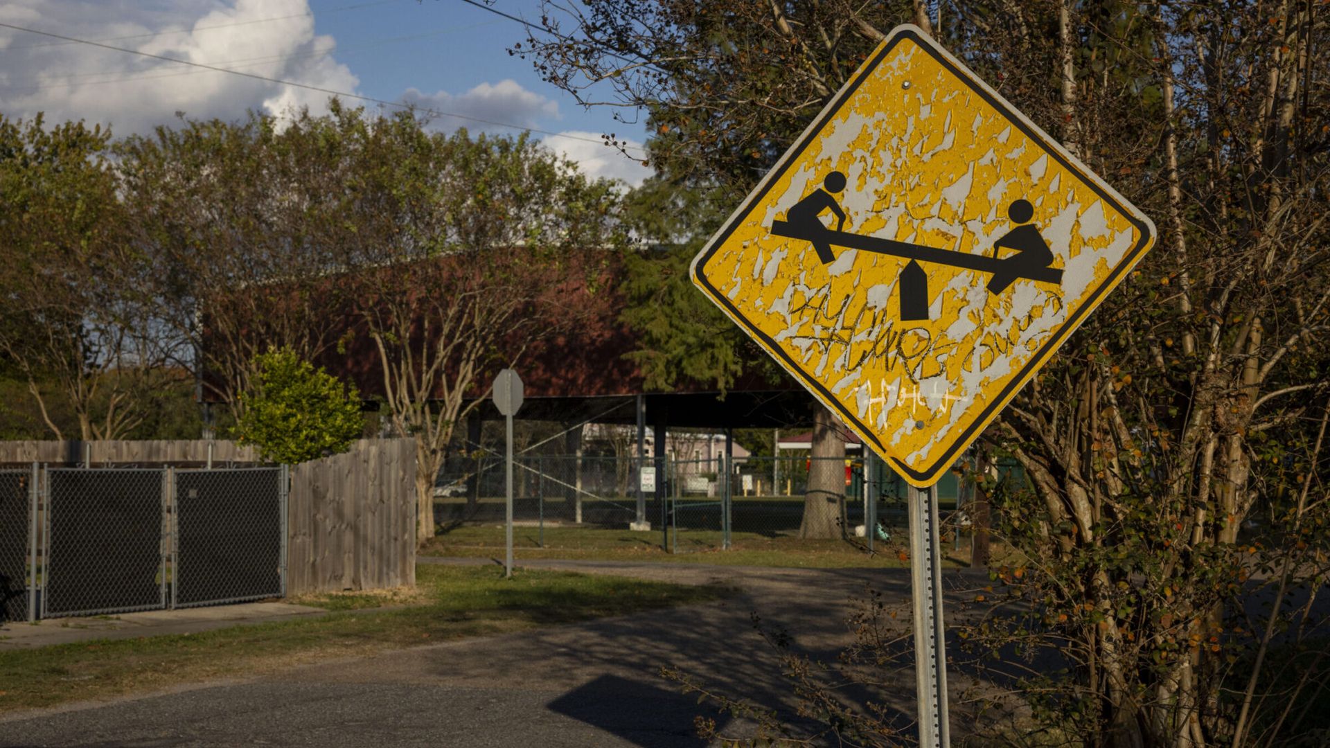 Weathered yellow diamond warning sign showing two children on a see-saw; peeling paint. Background includes chain-link fence, trees, blue sky with clouds, and a quiet street corner.