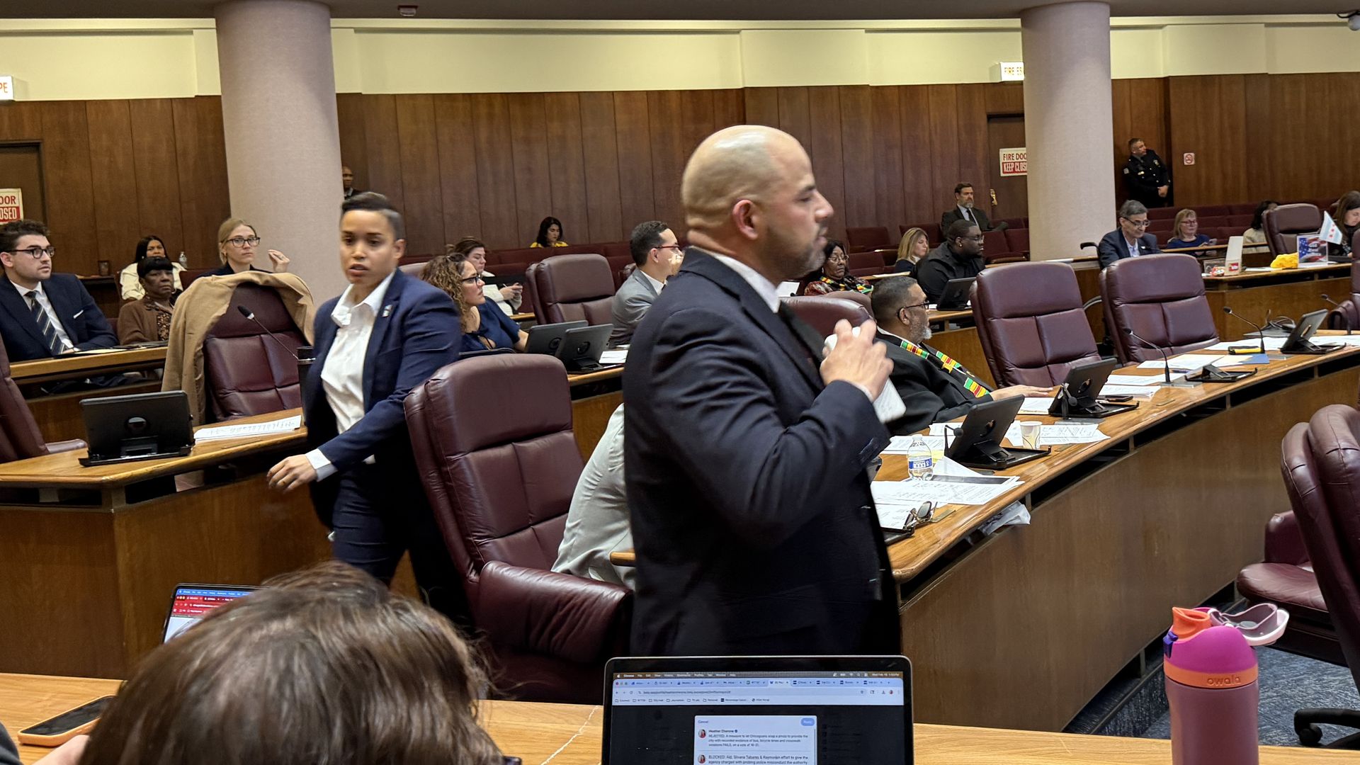People in business attire in a meeting room with wooden desks and leather chairs, some seated and some standing, engaging in discussion or working on laptops.