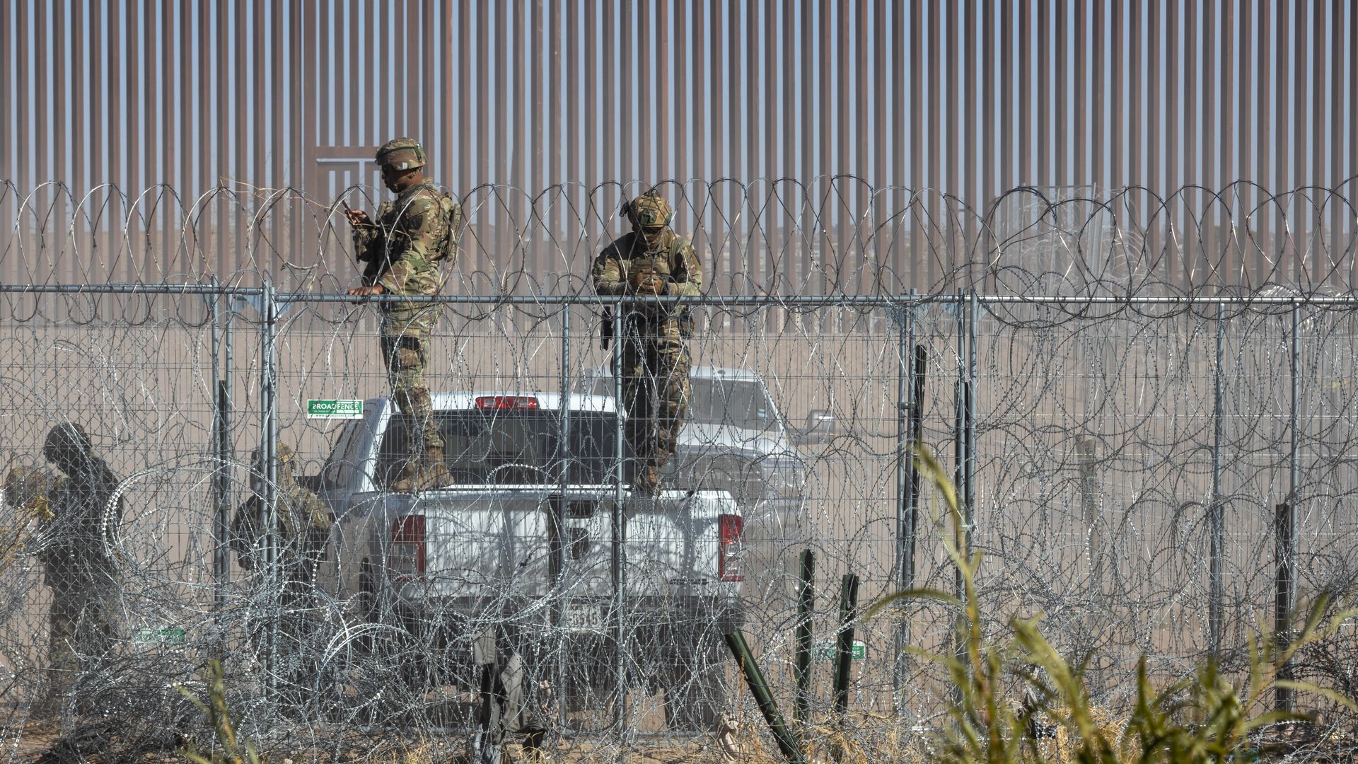 Two troopers with helmets and green camouflage stand on the back of a white truck in front of the border wall, with razor wire and a chain link fence in the foreground.